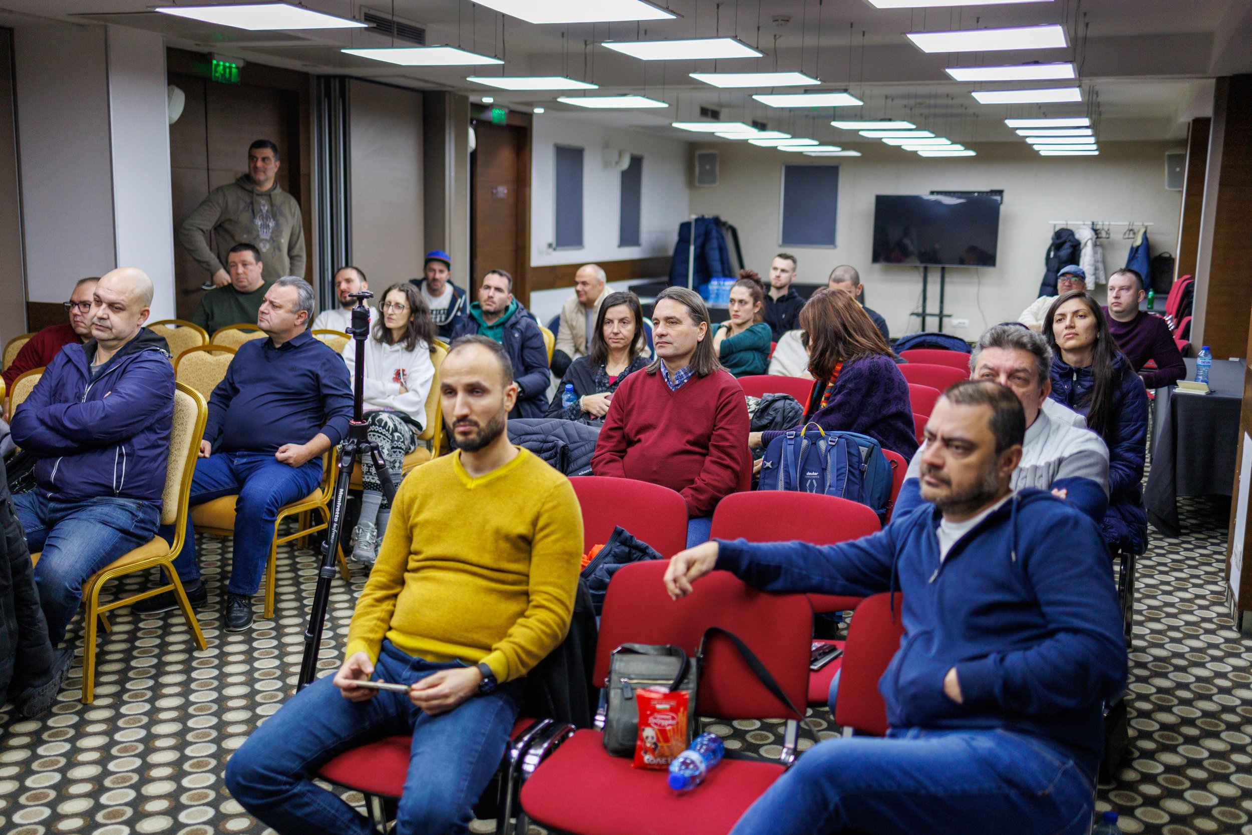 People attending a meeting or seminar in a conference room, seated in rows facing the front, with some taking notes or listening attentively.