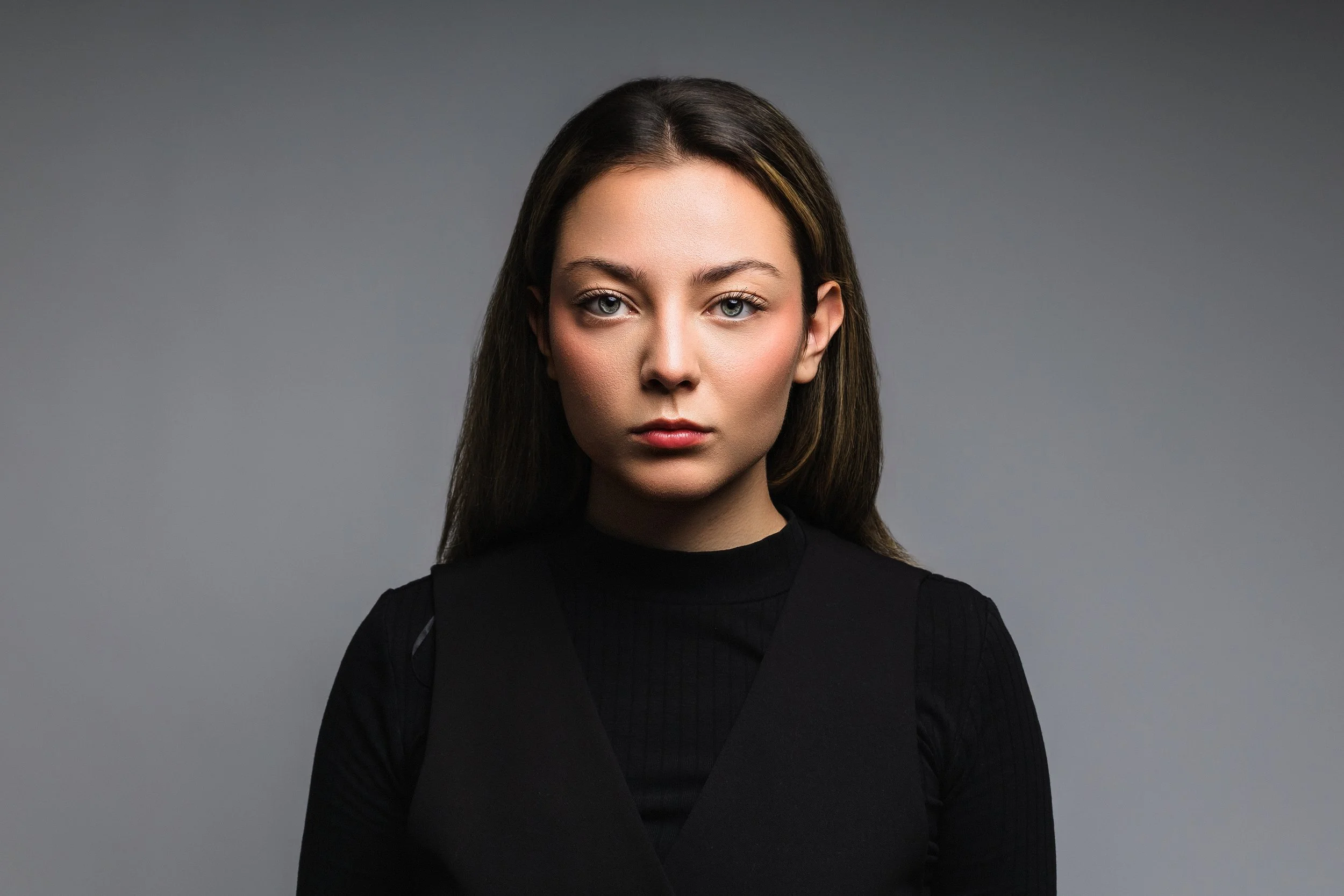Portrait of a young woman with straight brown hair, fair skin, blue eyes, dressed in black, against a gray background.