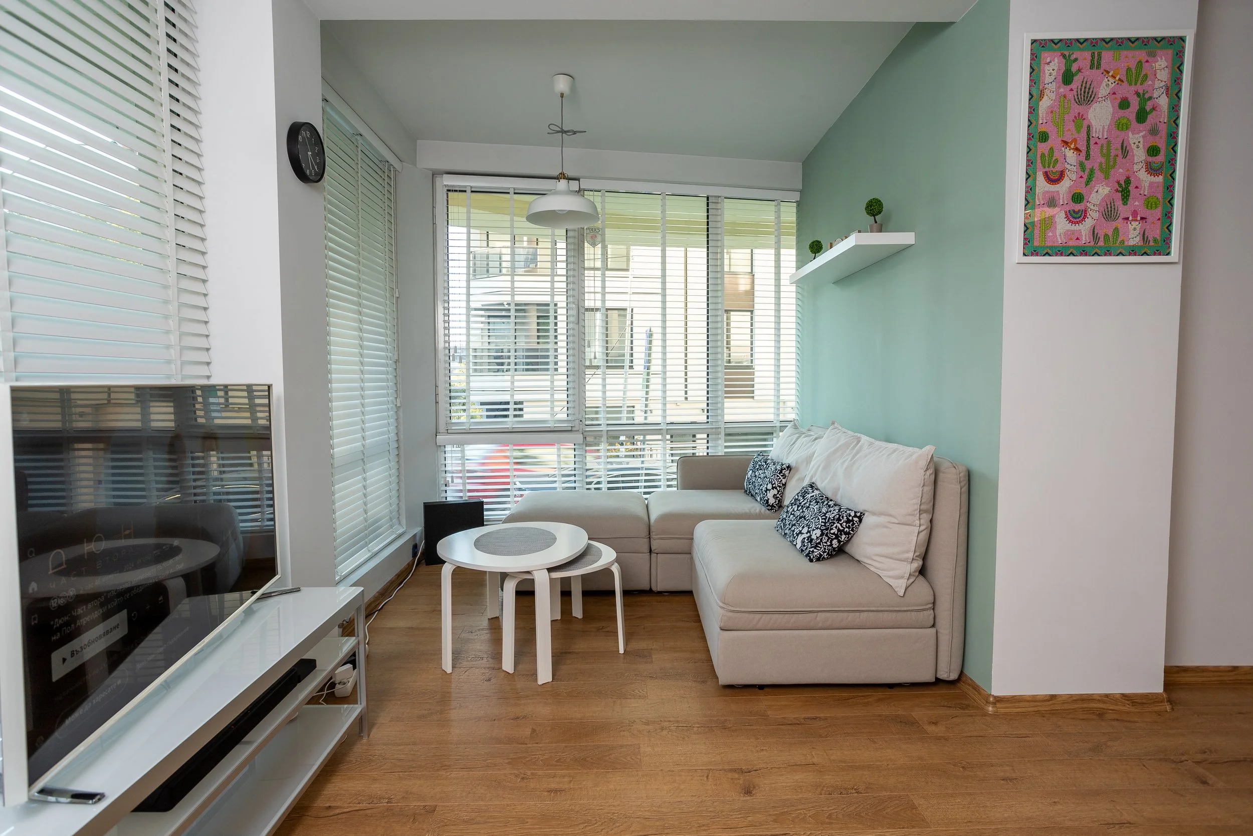 Living room with a beige sectional sofa, wooden floors, a white TV stand with a television, a small round coffee table, large windows with blinds, a teal accent wall with a colorful framed art piece, and some decorative plants on a white shelf.