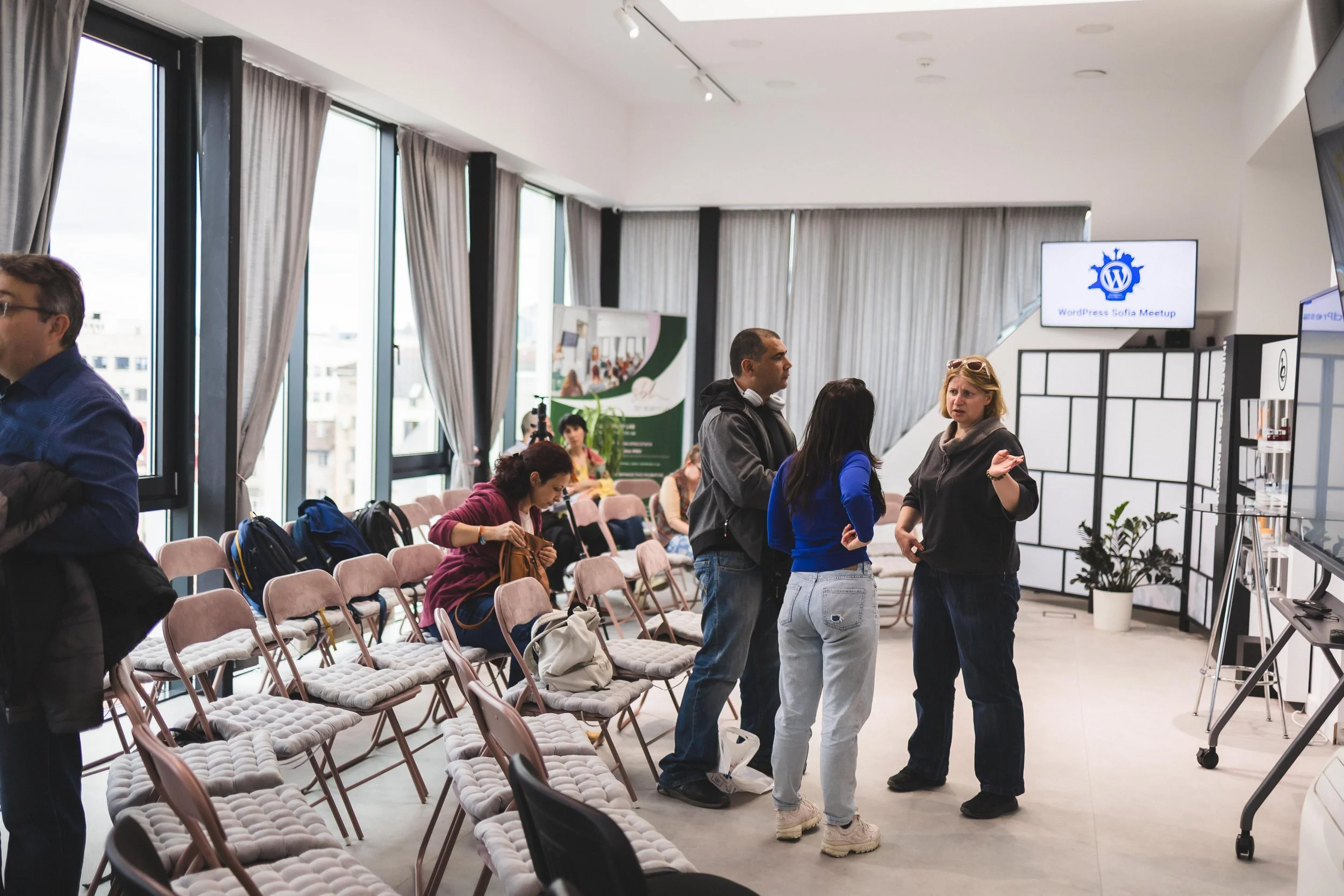People gathering in a modern conference room with rows of pink chairs, large windows, and a digital screen displaying 'WordPress Sofia Meetup', some are standing and chatting, others seated and preparing.
