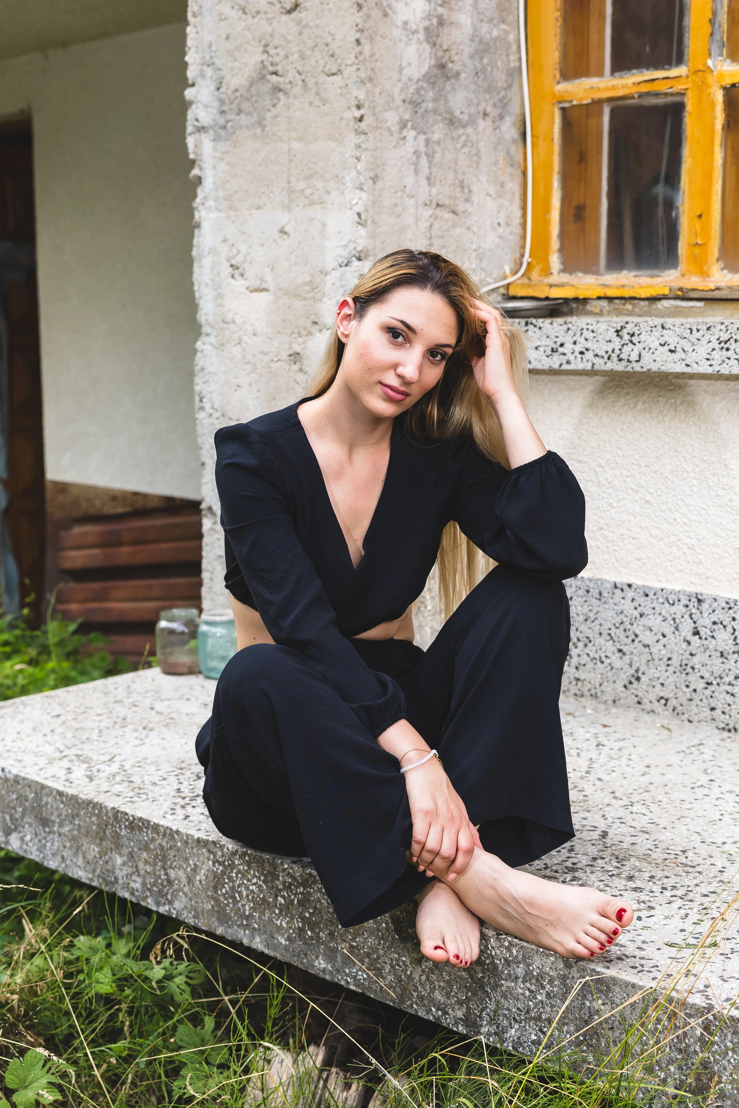 A young woman with long blonde hair, dressed in a black outfit, sitting barefoot on a concrete ledge outside an old building with a yellow window frame.