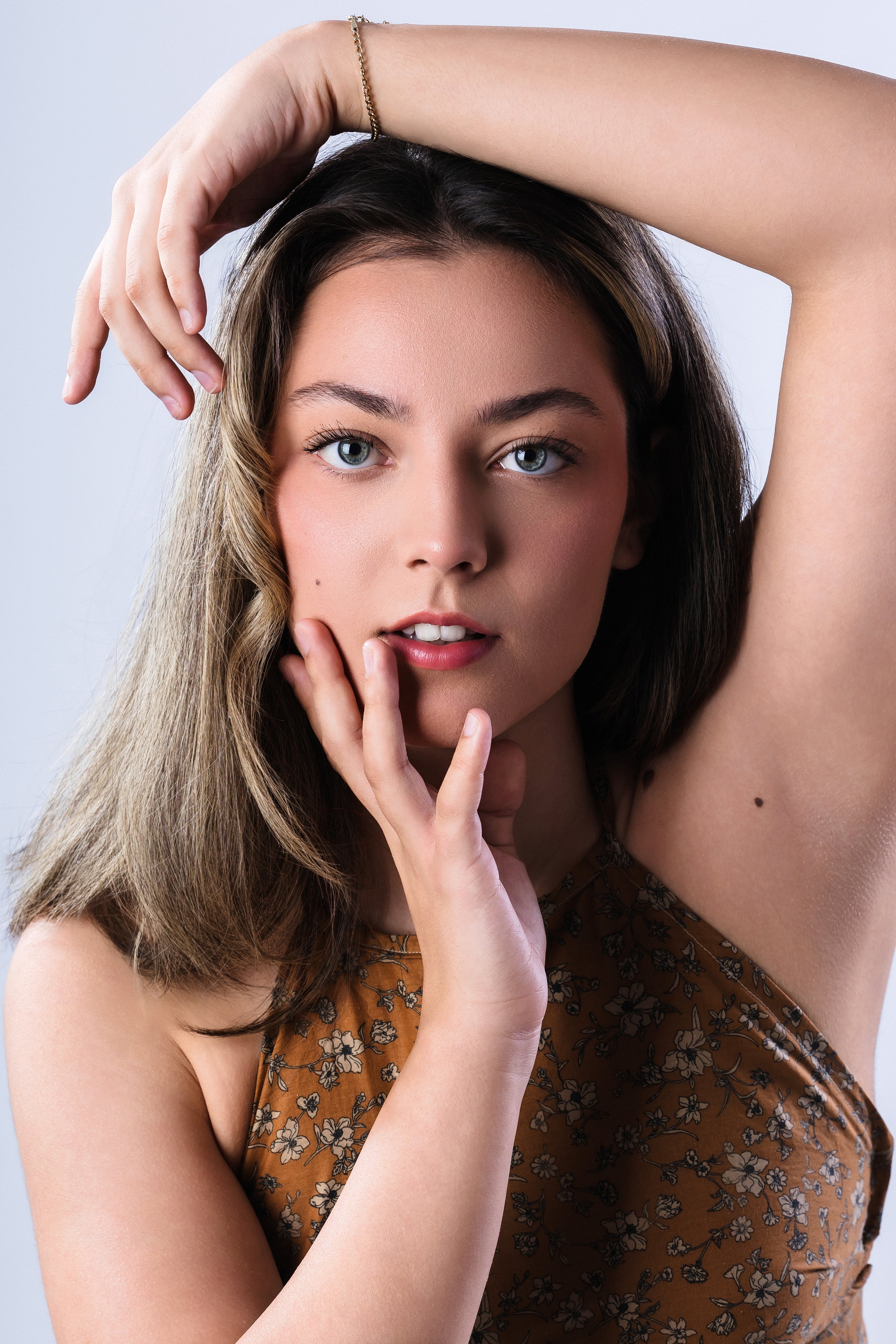 A young woman with blue eyes, long brown hair, wearing a floral brown sleeveless top, with one hand touching her face and the other raised above her head.
