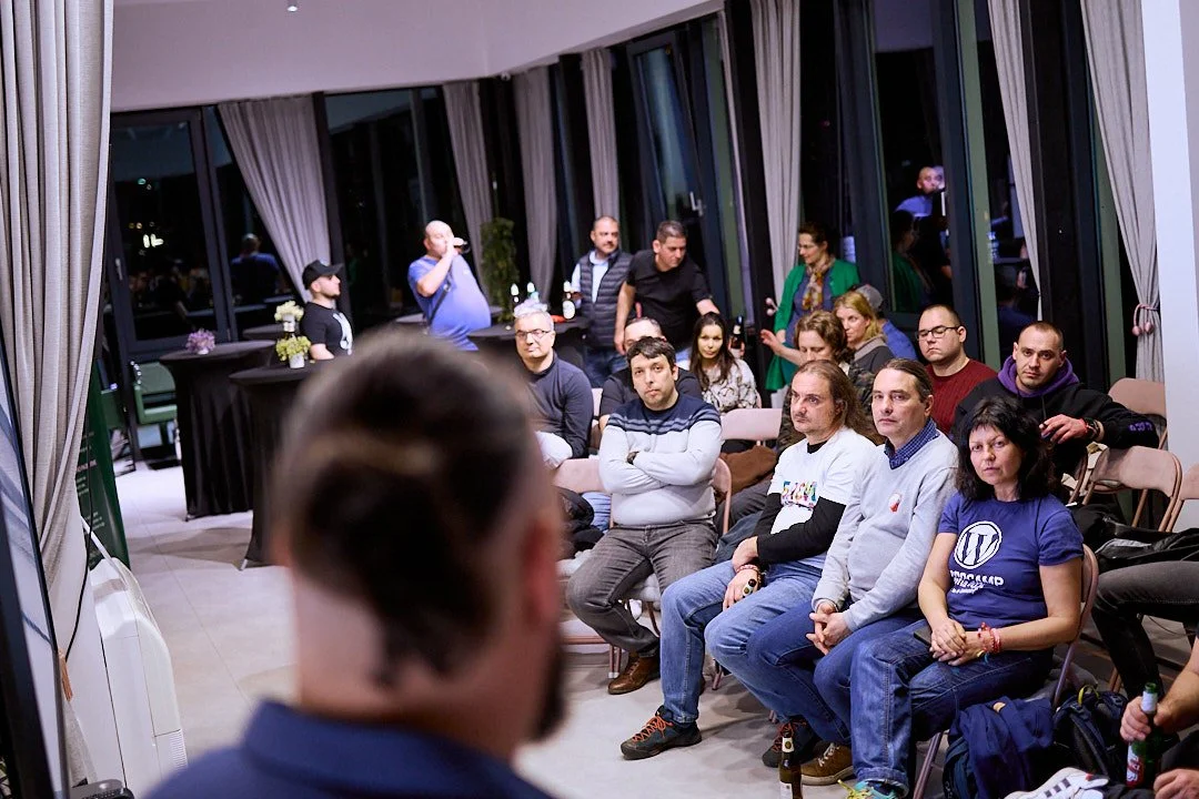 People attending a presentation or event in a large room with floor-to-ceiling windows, seated and standing, with a man in blue speaking or preparing to speak in the foreground.