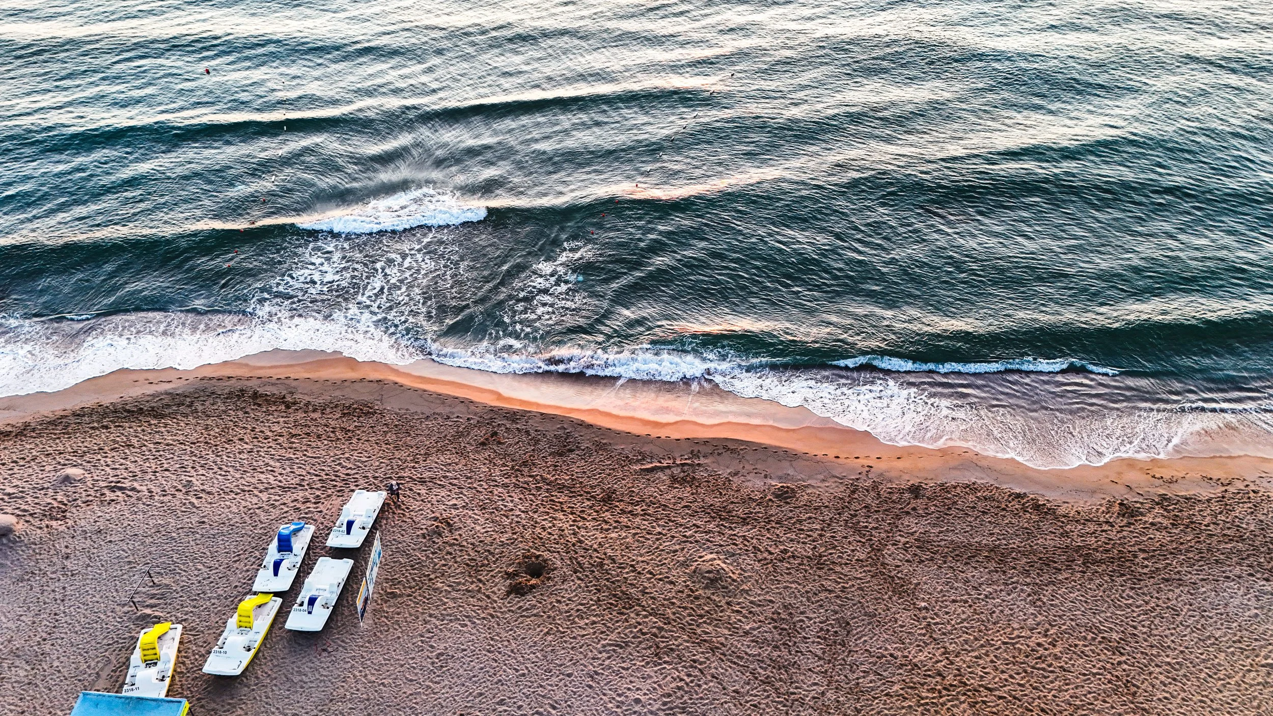 View of a beach with sand, small waves in the ocean, and several paddle boats lined up on the sand.