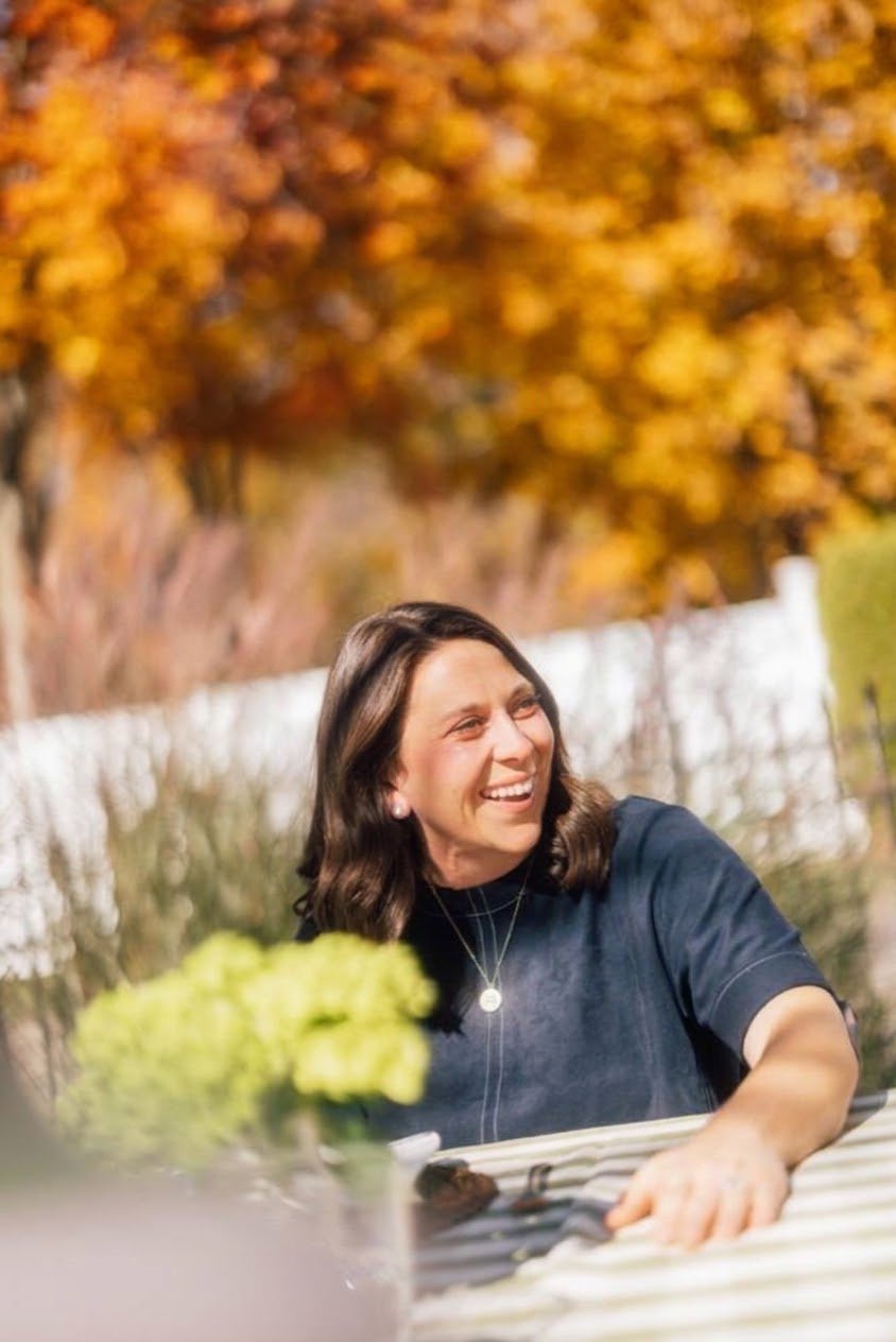 Woman smiling outdoors with a table set, under a partly cloudy blue sky.