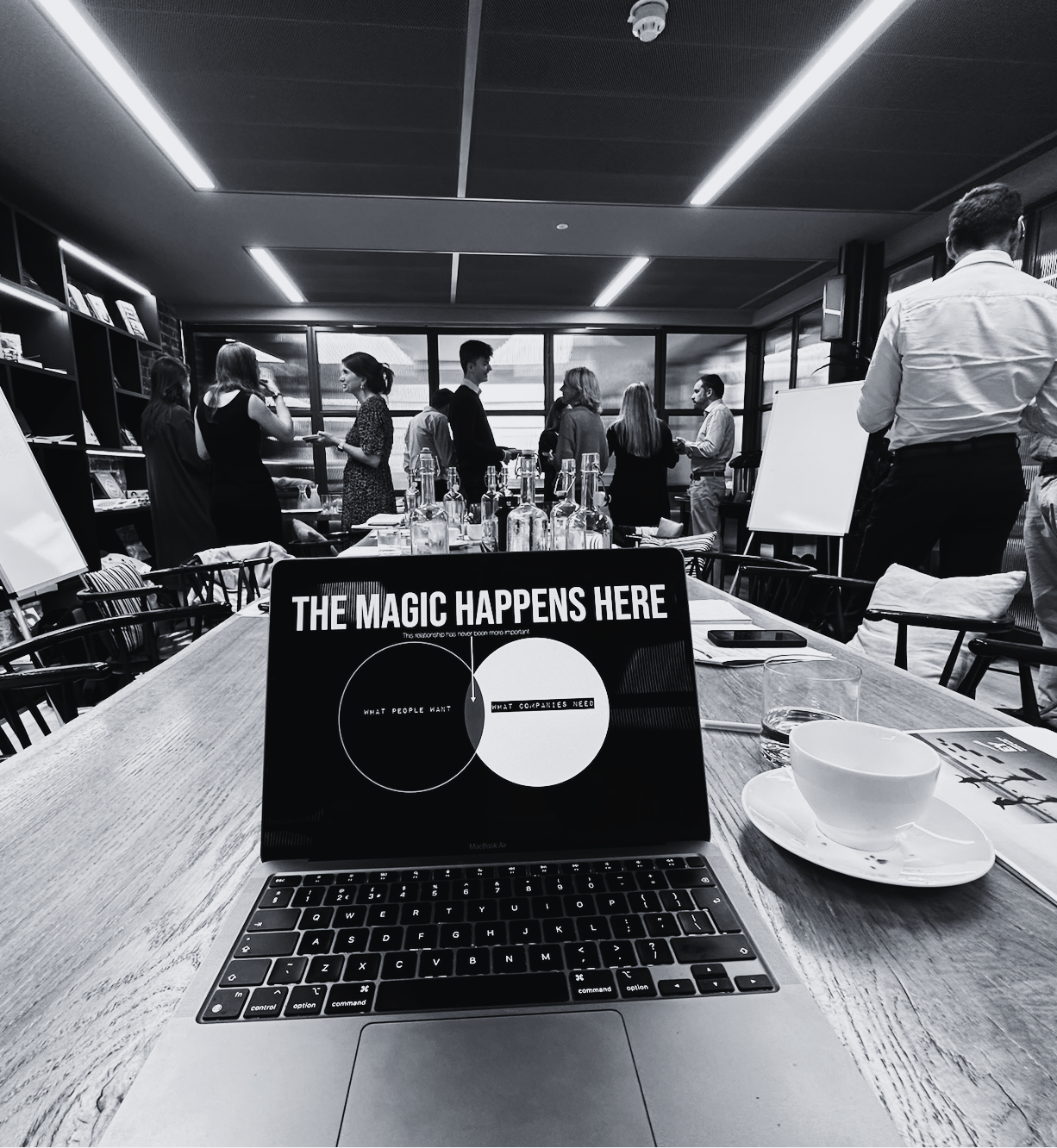 A conference room with people standing and talking in the background. On the table in the foreground, a laptop displays a slide with the title 'THE MAGIC HAPPENS HERE' and two overlapping circles labeled 'What People Want' and 'What Companies Need.' There is also a cup on a saucer, a glass of water, a smartphone, and a pen on the table.