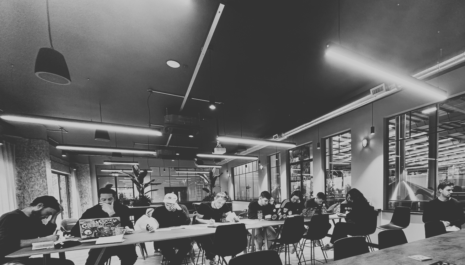 People working and studying at a long table in a modern cafe or coworking space with large windows and industrial decor.
