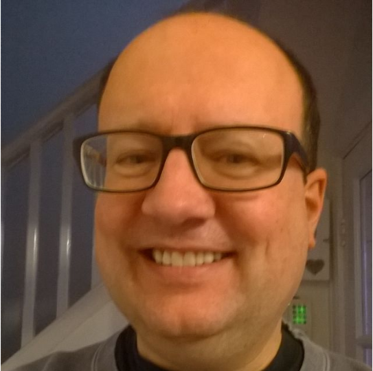 A close-up photo of a smiling man with glasses, short dark hair, and light skin, indoors with a window and some household objects in the background.