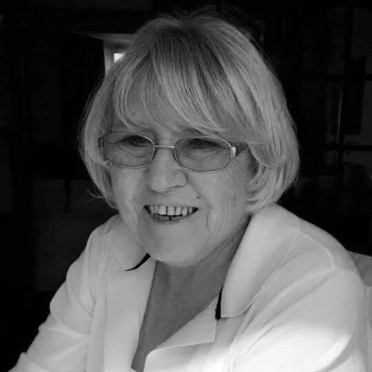 A smiling woman with short blonde hair, glasses, and a white blouse sitting indoors.