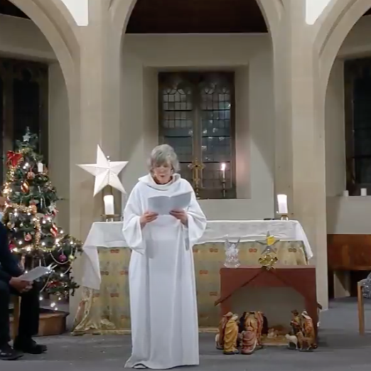 An elderly woman dressed as an angel, wearing a white robe, is standing and reading from a book in a church decorated for Christmas. There is a Christmas tree decorated with ornaments and lights to her left, and a nativity scene with figurines to her right. The church interior features arched windows and warm lighting.