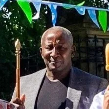 A man in a gray suit holding a ceremonial staff, standing outdoors with trees and decorations in the background.