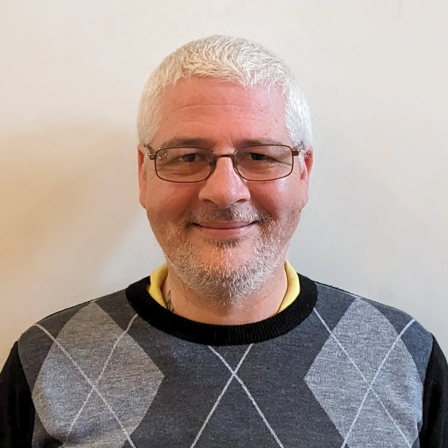 A man with white hair and beard wearing glasses and a grey argyle sweater, smiling, against a plain beige background.