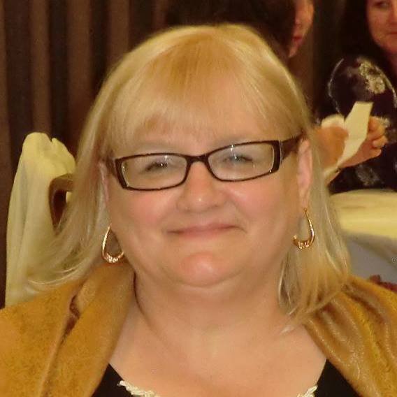 A smiling woman with light blonde hair, glasses, and gold earrings sitting at a table in a restaurant.