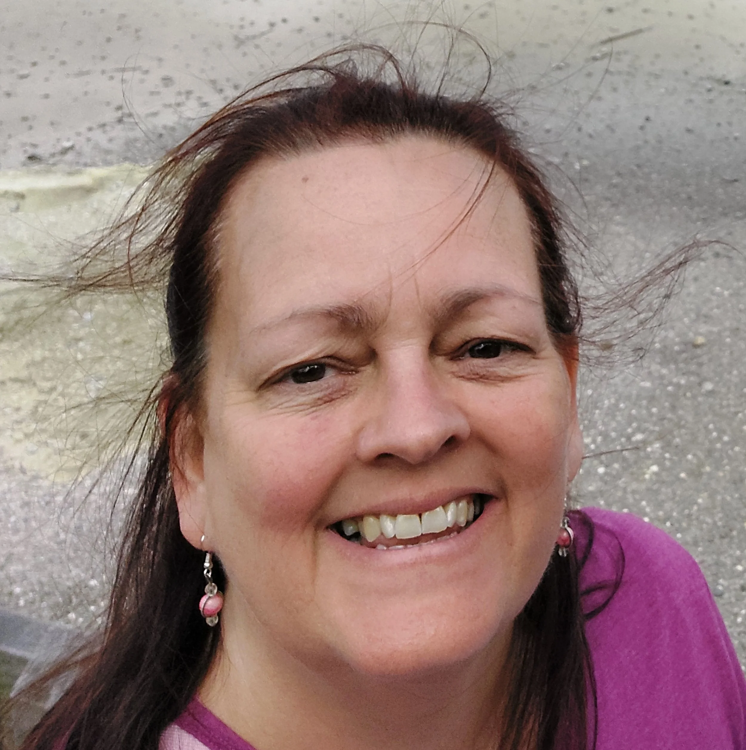 Close-up of a smiling woman with brown hair and earrings, outdoors on a windy day.