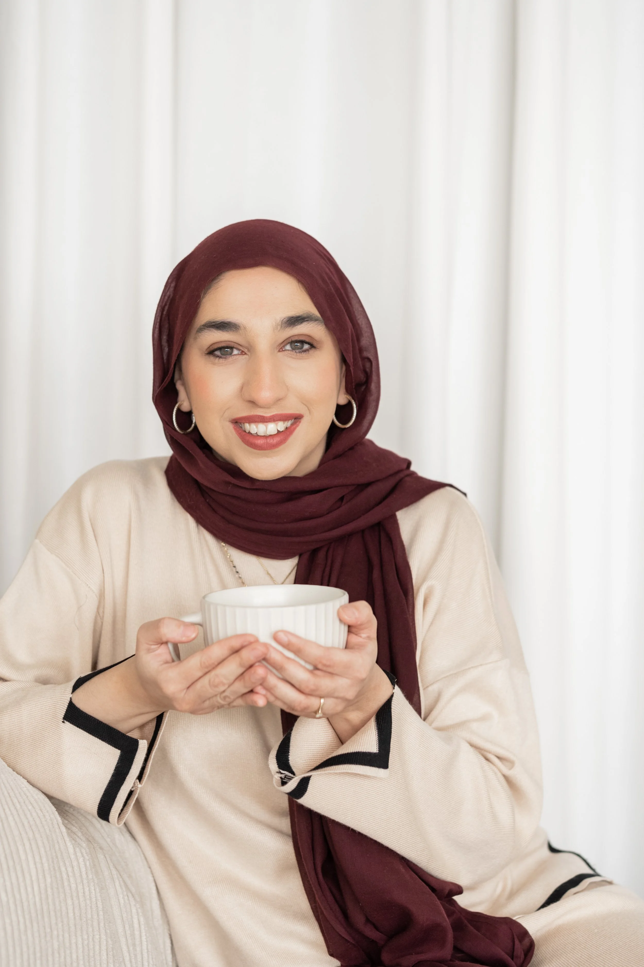 A woman wearing a maroon hijab and beige outfit smiling while holding a white cup, sitting in front of a white curtain.