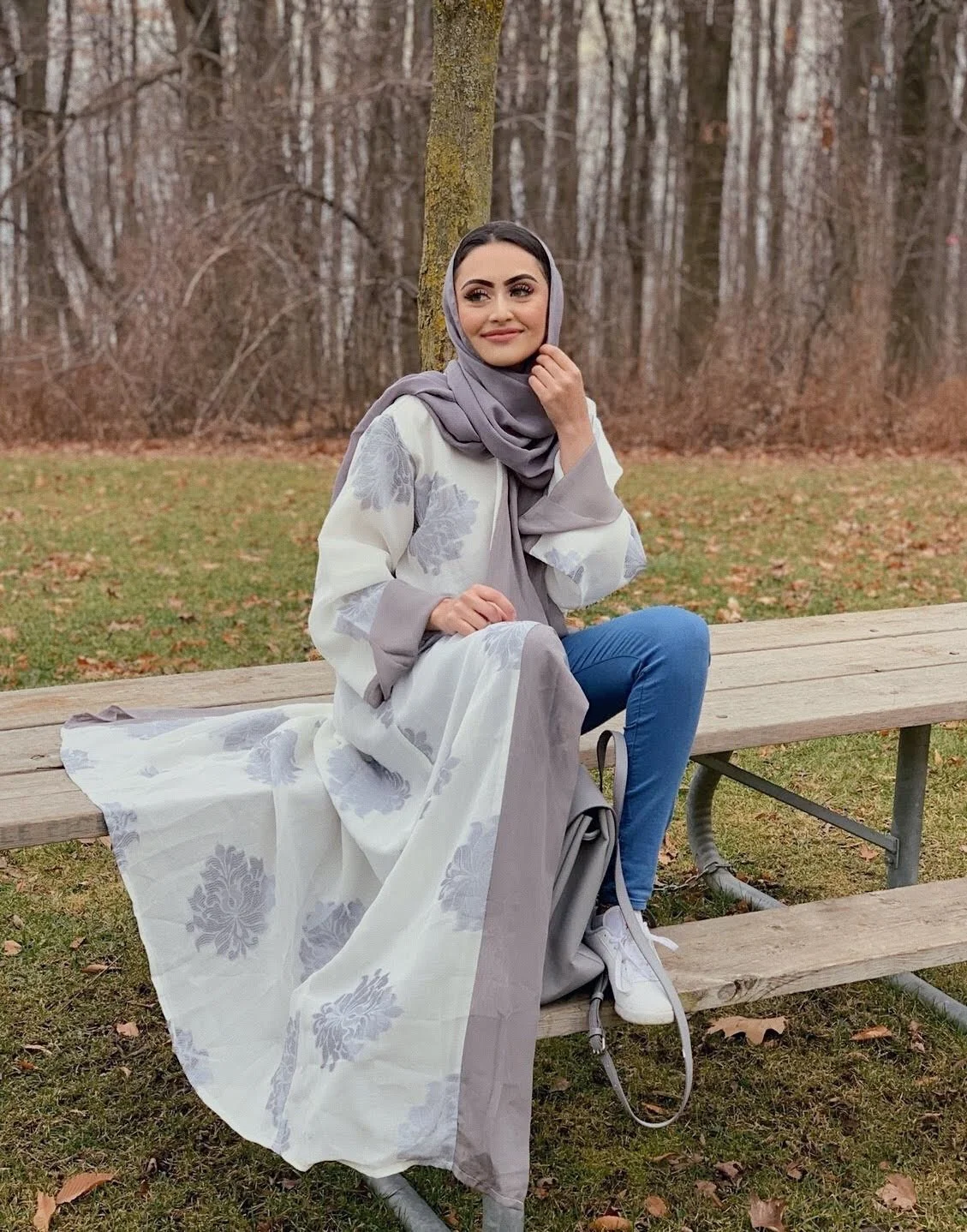 A woman seated on a wooden bench in a forest. She is wearing a long light-colored dress with floral patterns and a matching headscarf, blue jeans, and white sneakers. She is smiling with her hand resting on her chin, surrounded by bare trees and fallen leaves.