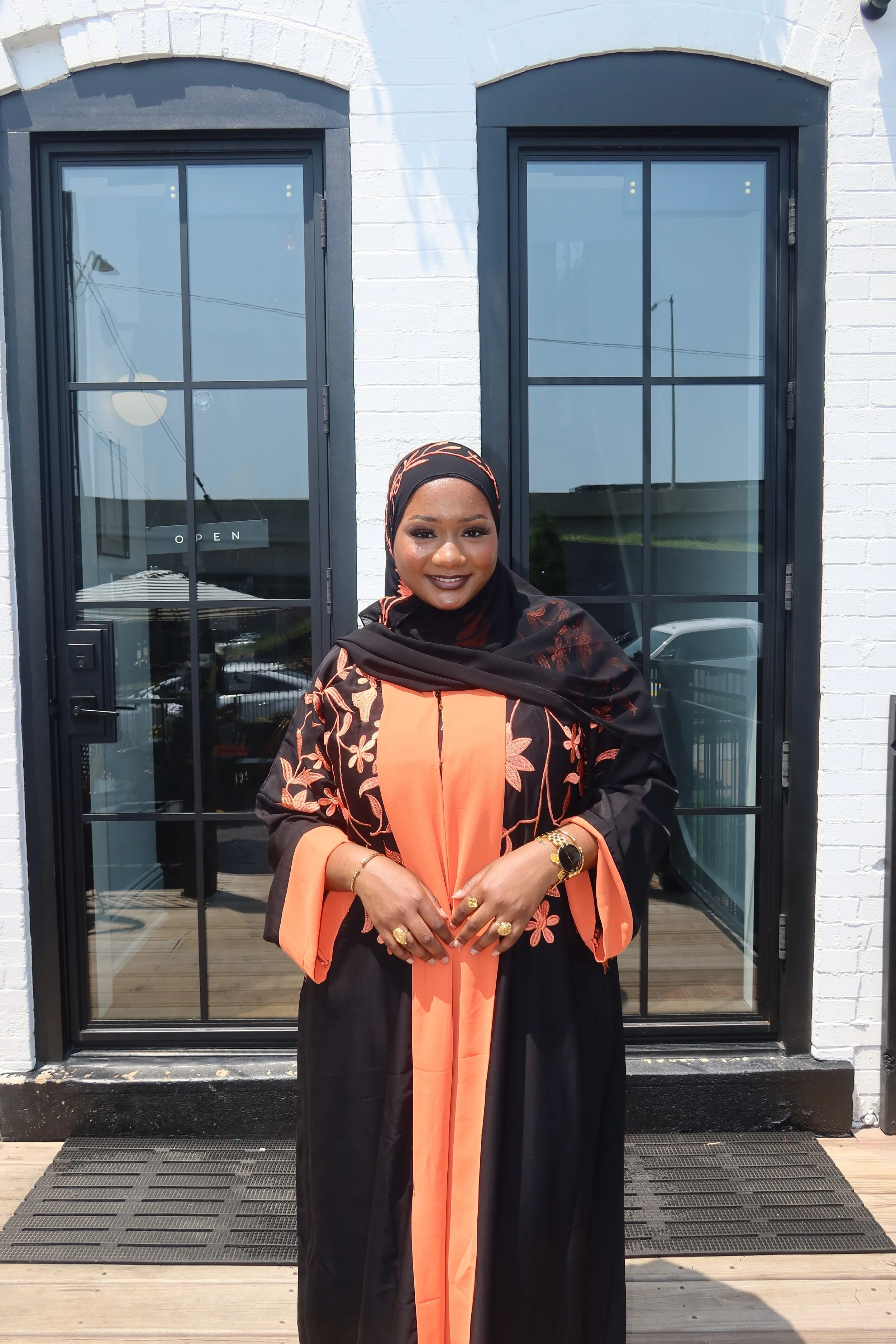 Woman wearing a black and orange floral dress and headscarf, standing in front of two glass doors.