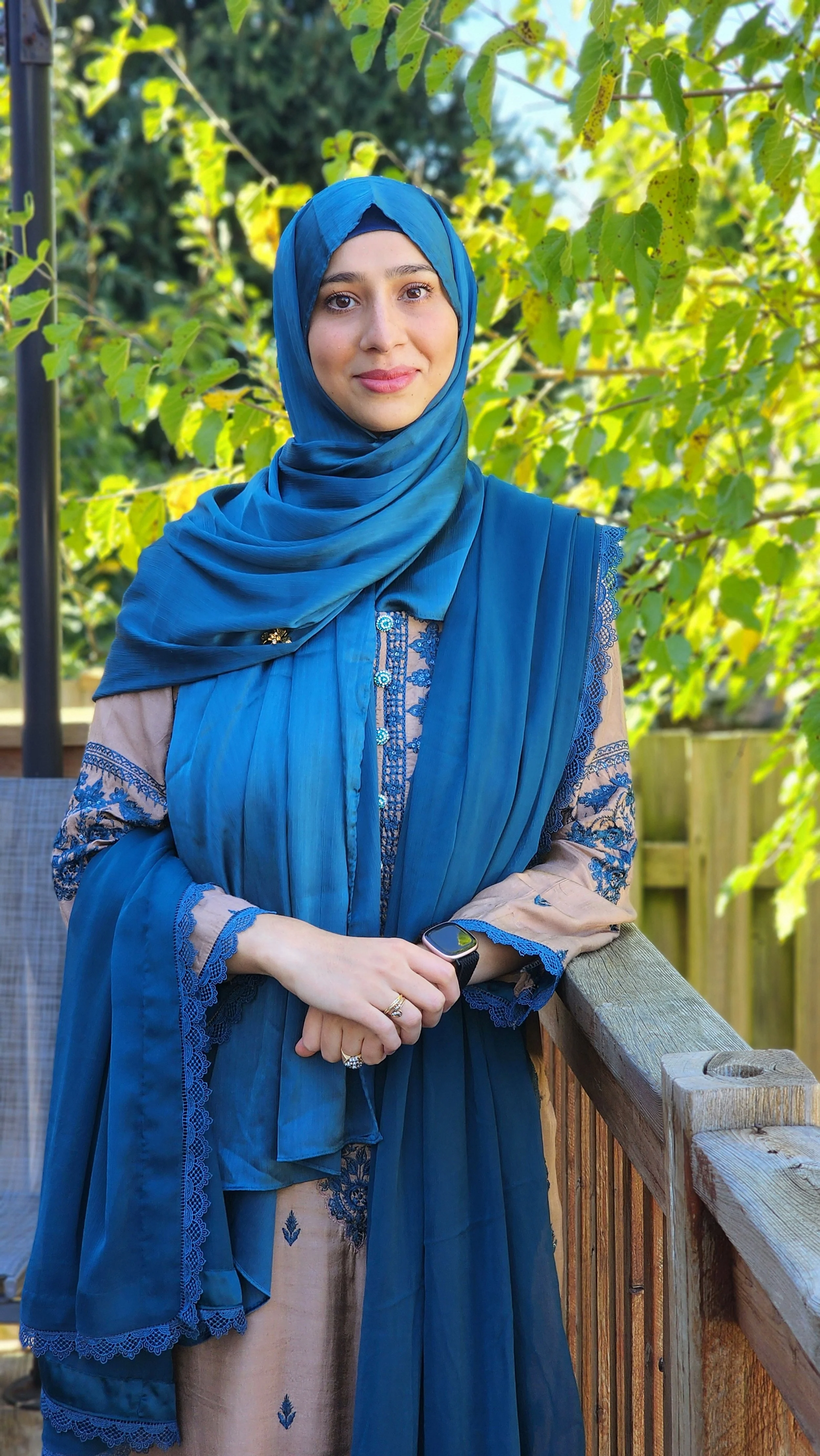 Smiling person in a blue hijab and traditional outfit on a balcony with leafy background