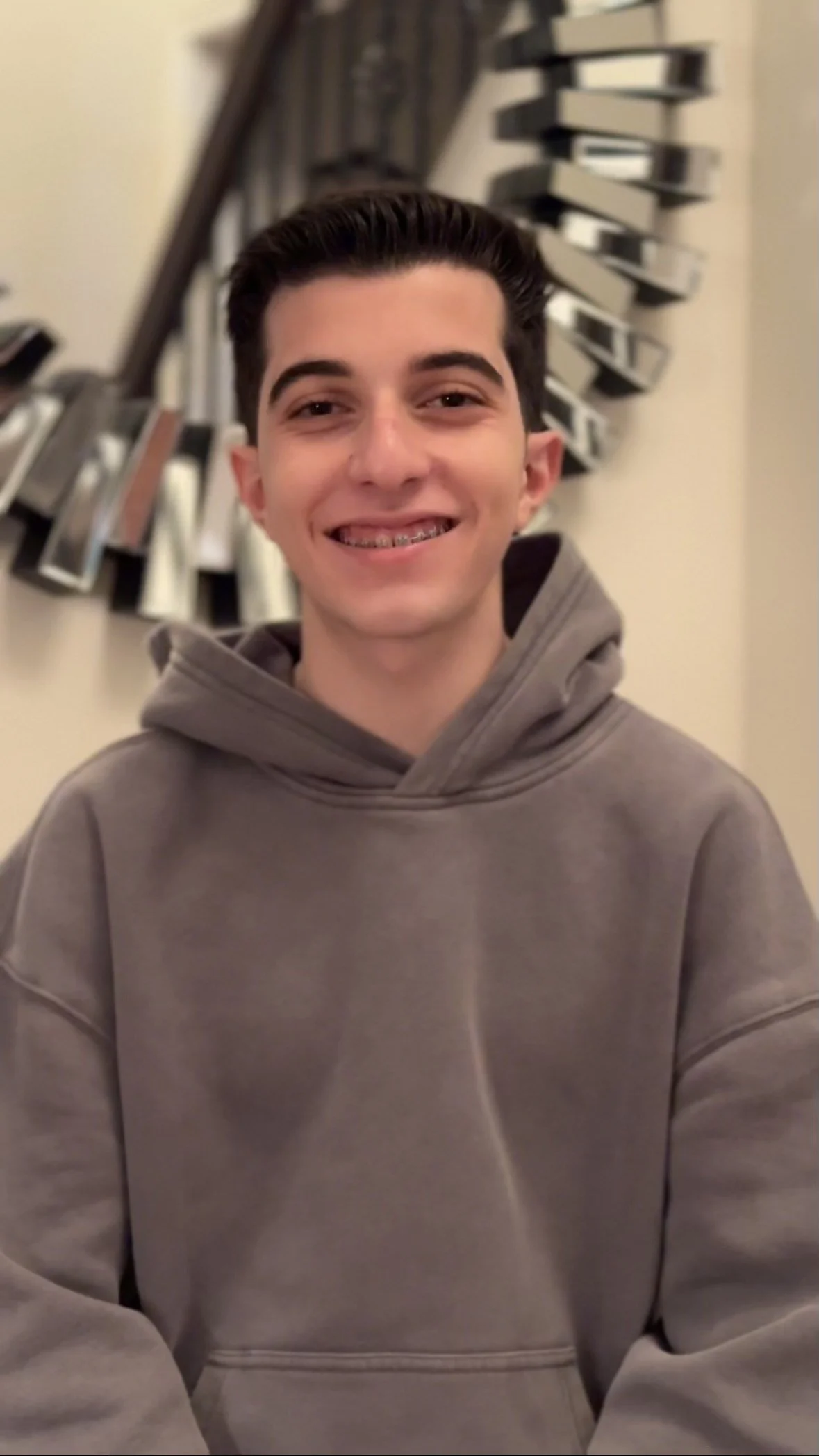 Person wearing a gray hoodie, smiling, with braces, standing indoors in front of a decorative mirror.