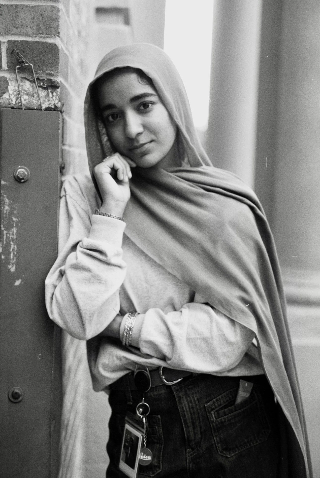 Black and white portrait of a person wearing a headscarf and casual clothing, leaning against a brick wall.
