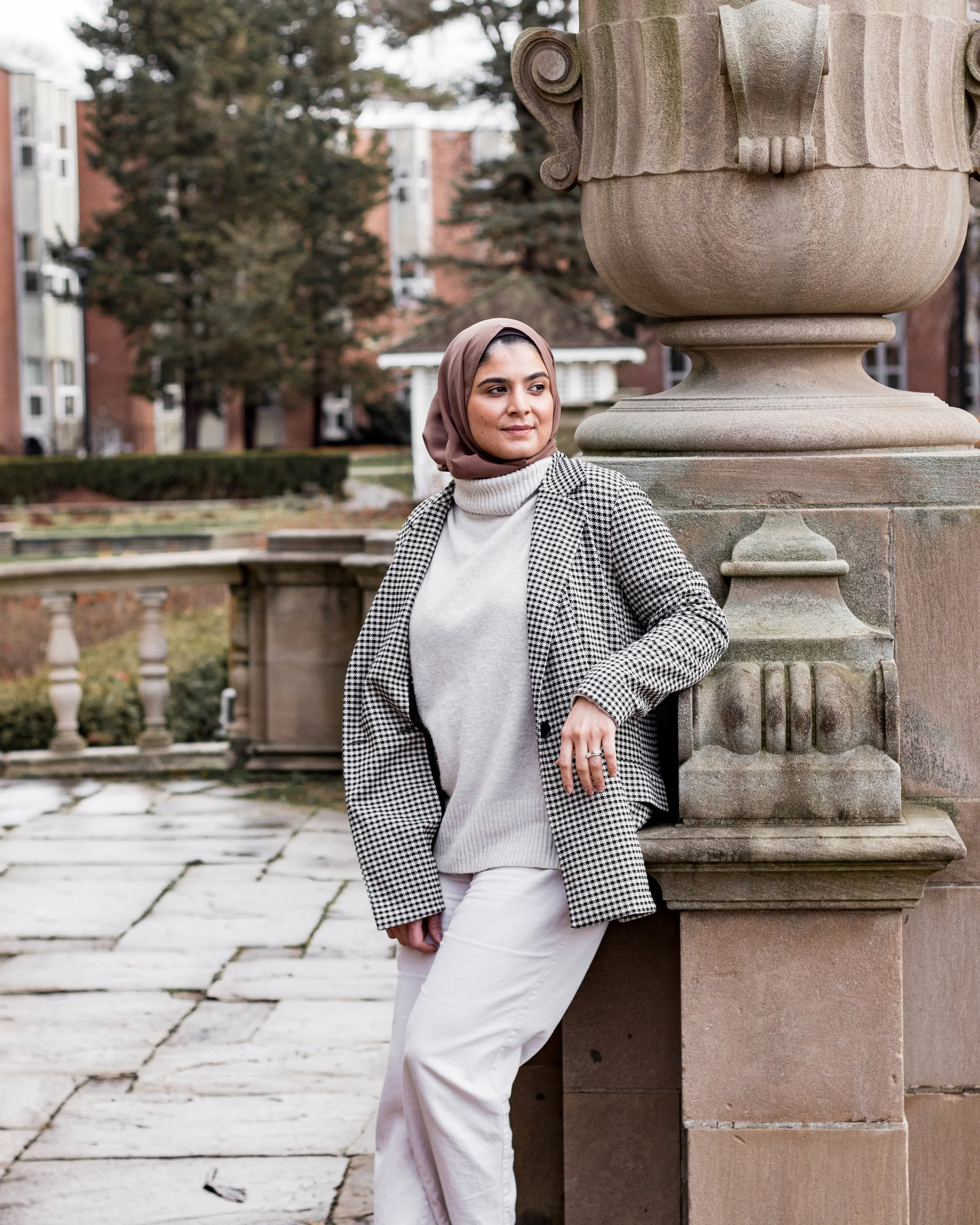 Woman wearing a hijab and houndstooth coat leaning against a stone column in an outdoor setting with trees and buildings in the background.