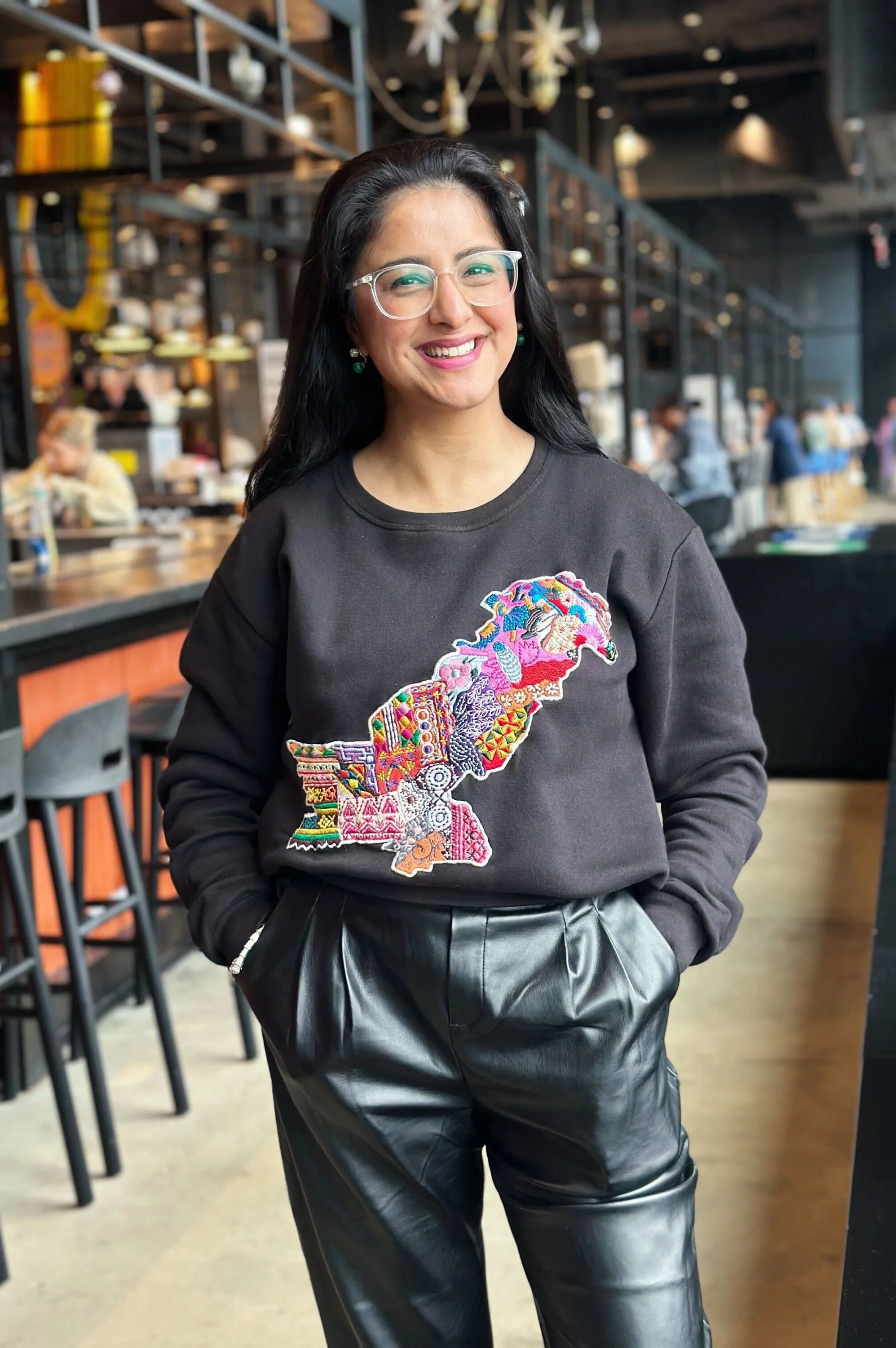 A woman smiling, wearing glasses and a black sweatshirt with colorful embroidery, standing in a modern café or restaurant with industrial design.