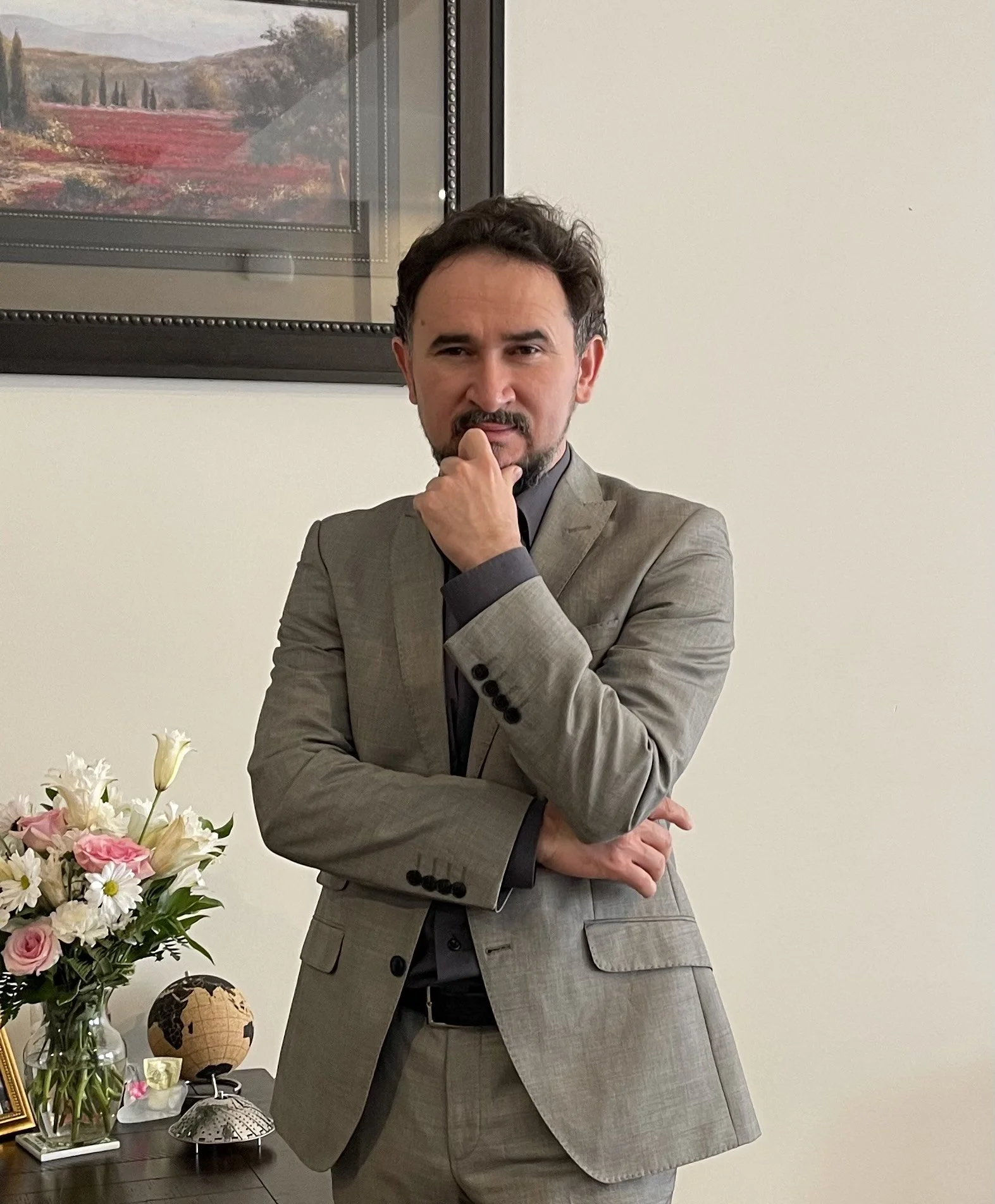 Man in gray suit posing thoughtfully with hand on chin near desk with flowers and globe.