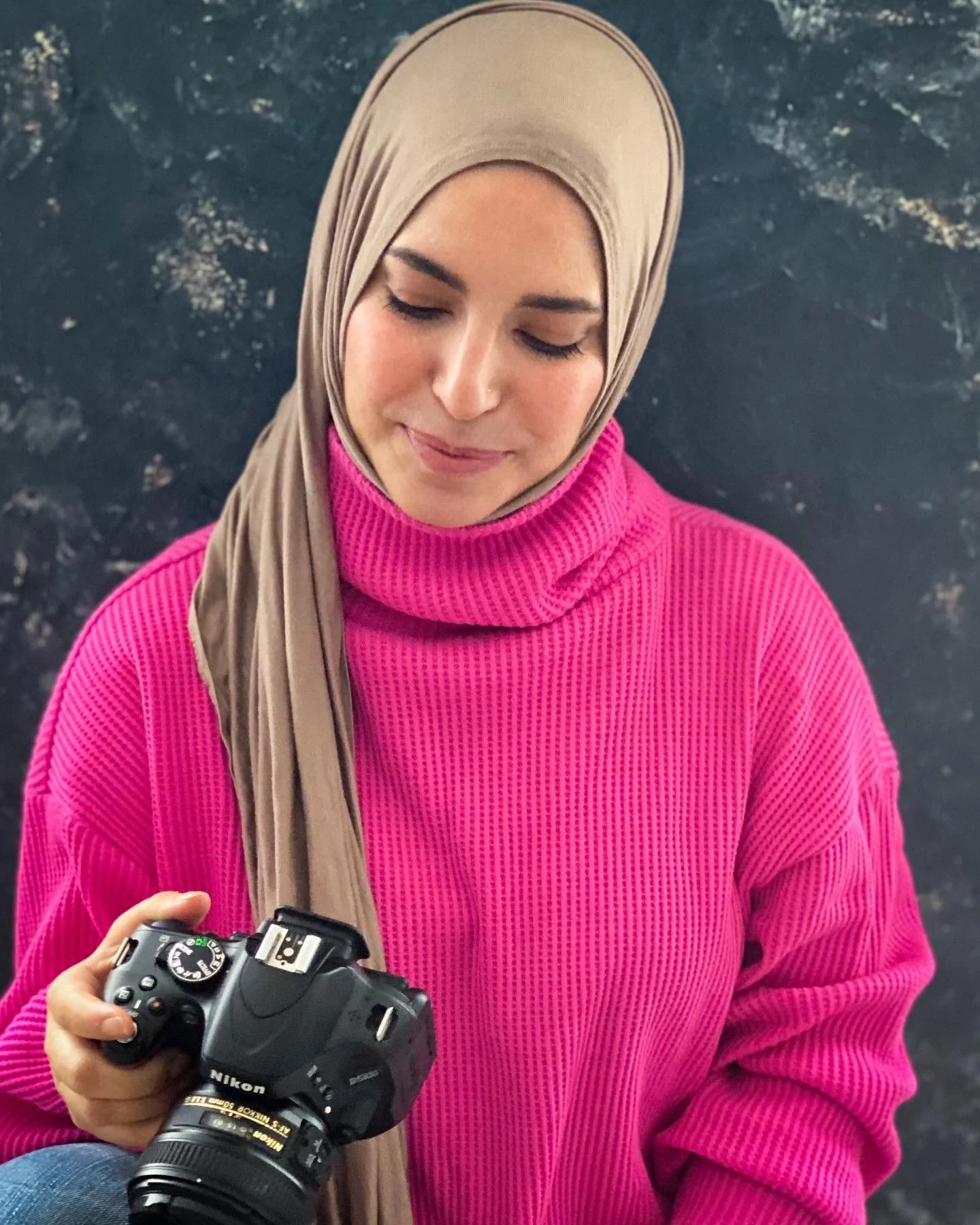A woman wearing a beige hijab and bright pink sweater, holding a Nikon DSLR camera.