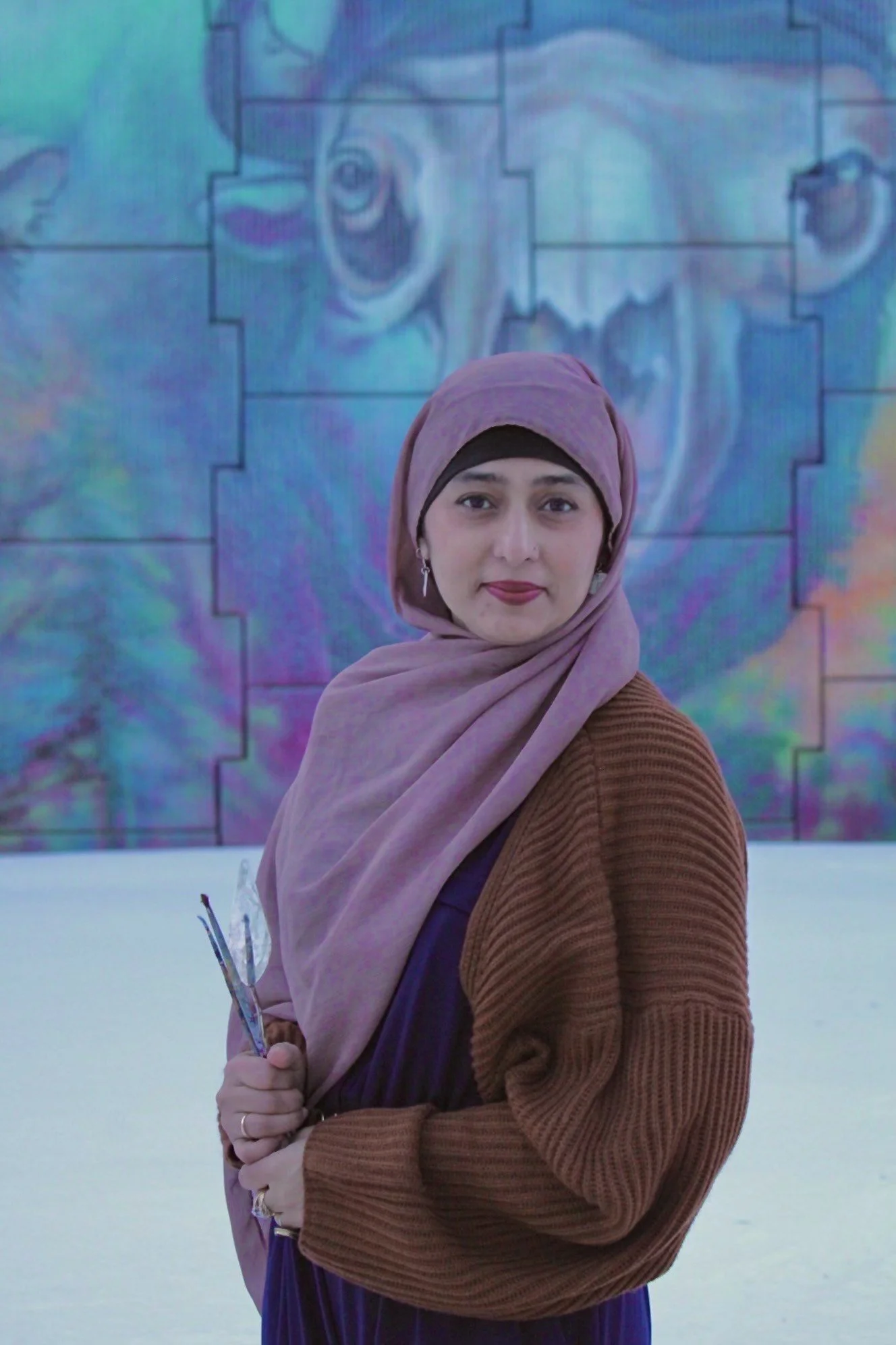 Woman in a hijab holding a paintbrush, standing in front of a colorful mural featuring an animal face.