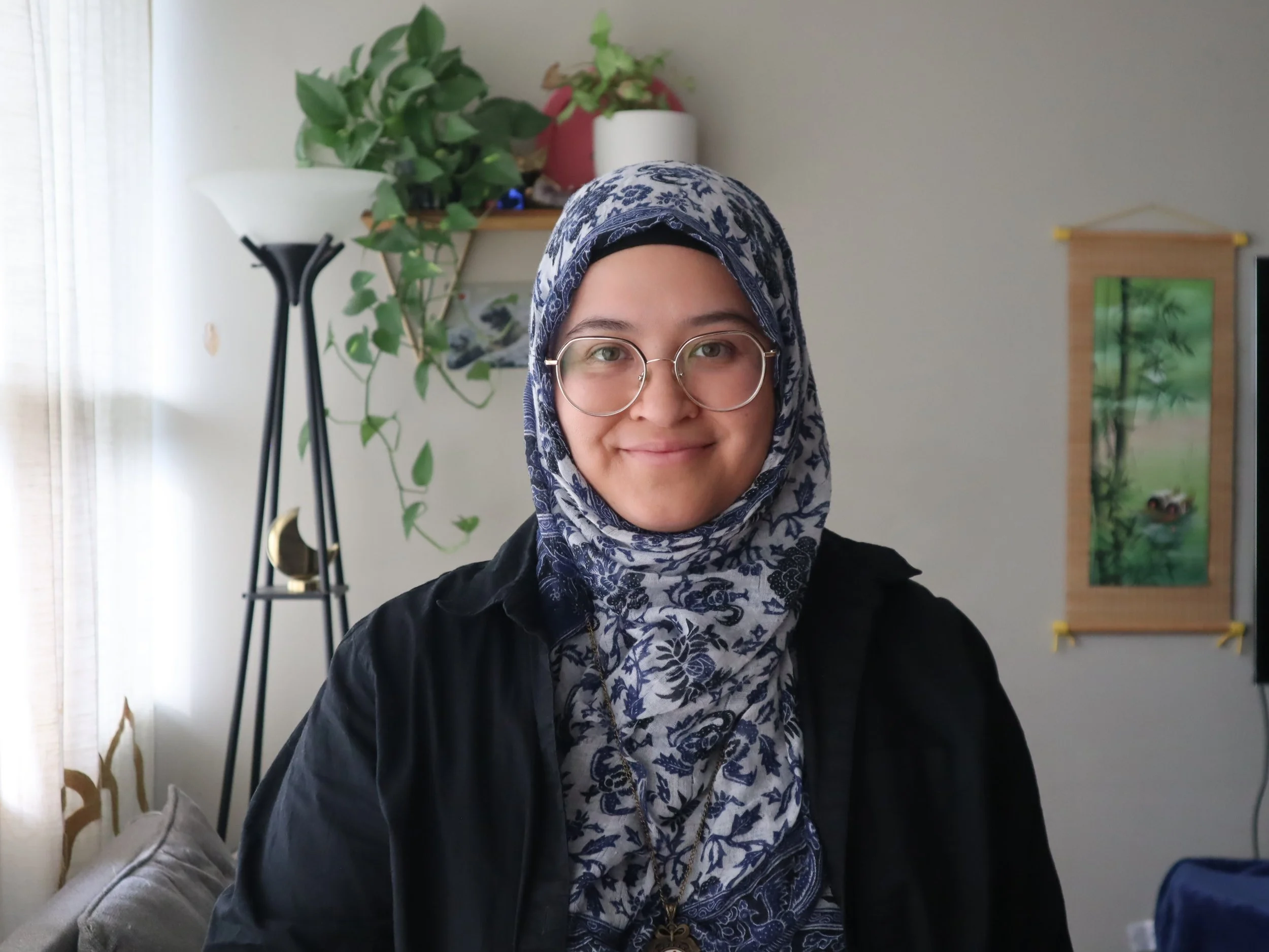 Person wearing a patterned hijab and glasses, smiling in a room with plants and decor.
