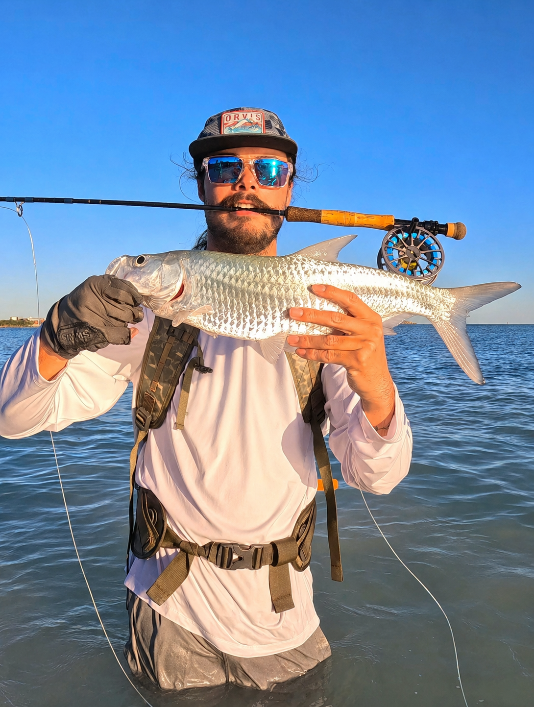 A man holding a large fish he caught while fishing in the water, wearing a cap, sunglasses, and a fishing harness, with a fishing rod in his mouth, on a clear day.