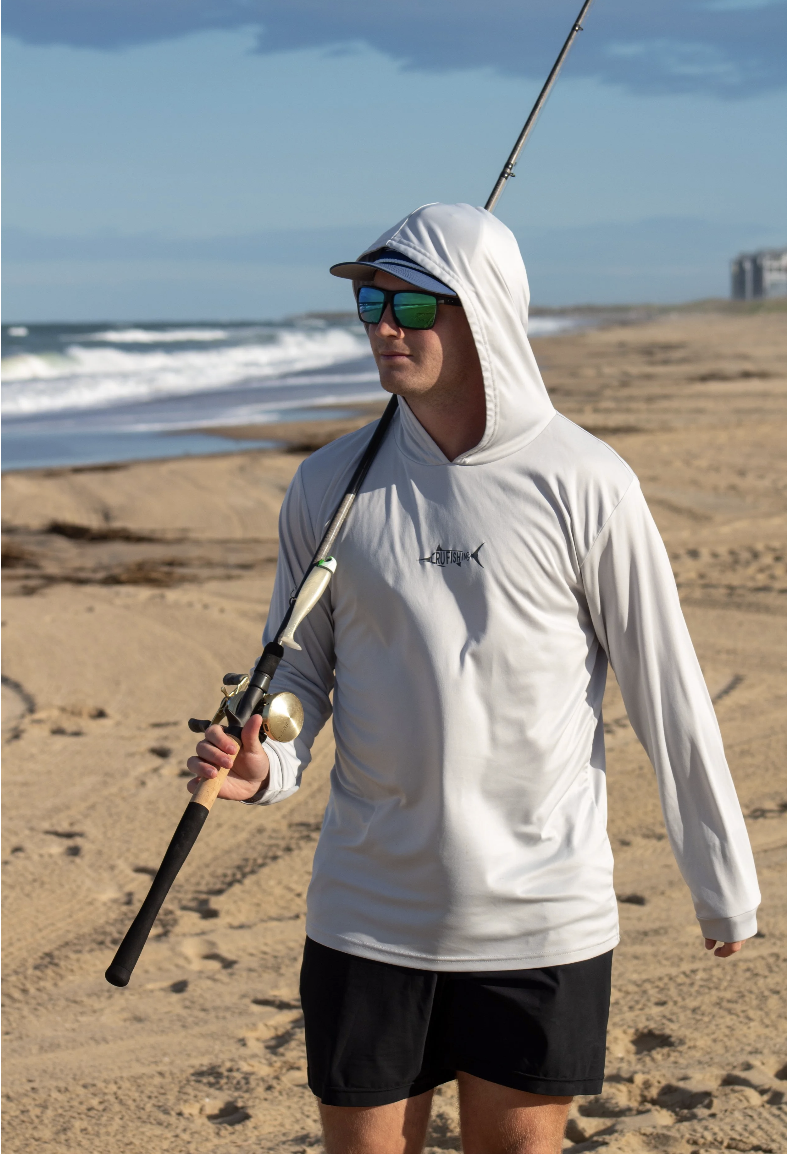 A man dressed in a white hoodie and black shorts walking on a sandy beach with a fishing rod.