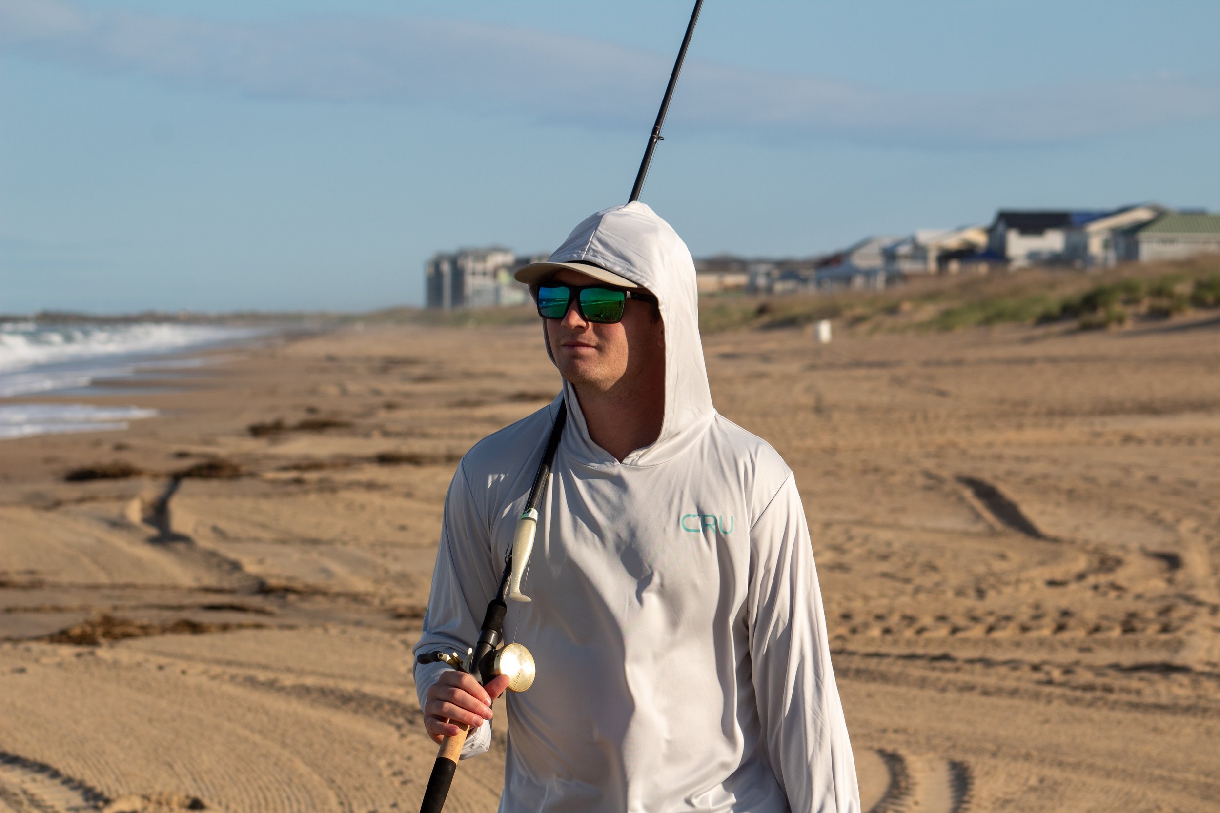 A man wearing a white hoodie, sunglasses, and holding a fishing rod stands on a sandy beach with houses in the background and ocean waves nearby.