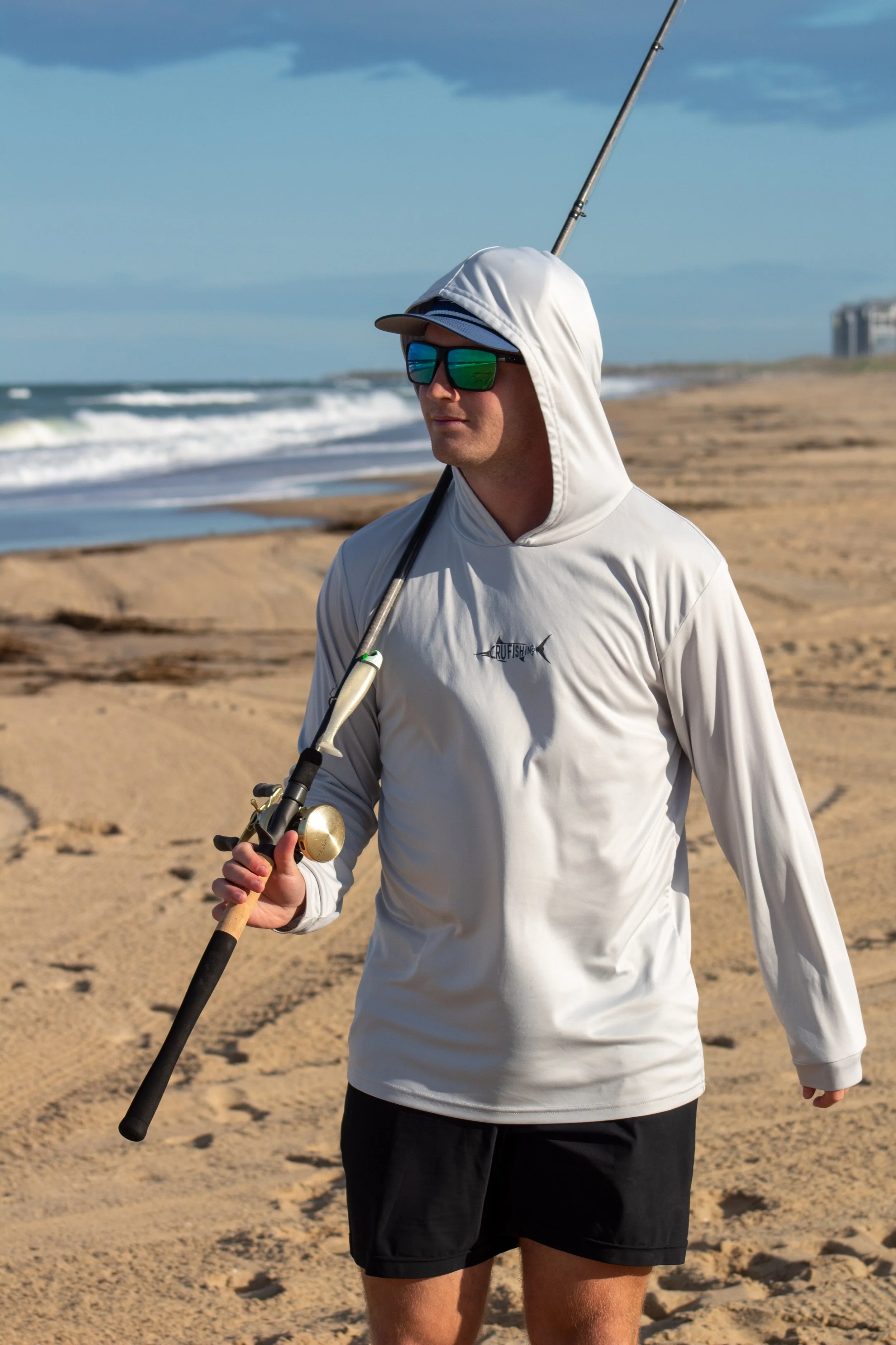 A man holding a fishing rod on the beach. He is wearing a white hoodie, sunglasses, and a cap, with the ocean and cloudy sky in the background.