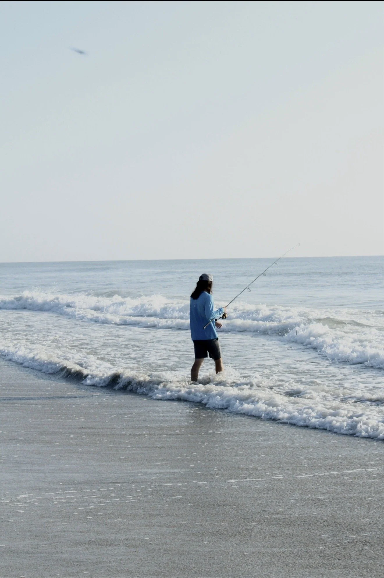 A person standing on the beach in the shallow water fishing with a rod and reel, wearing a blue jacket, black shorts, and a cap, with the ocean and a clear sky in the background.