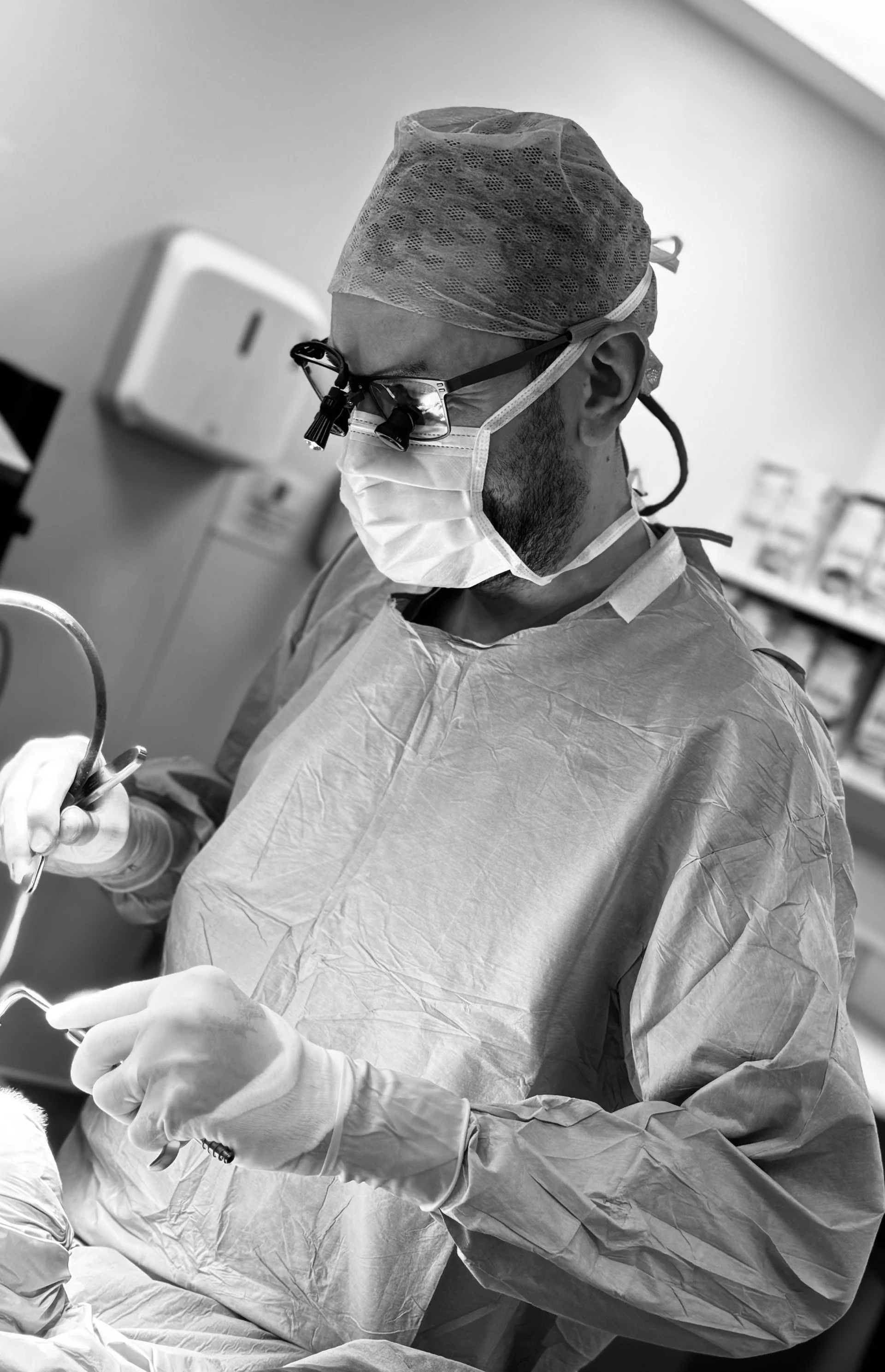 Mr Tim Biggs ENT surgeon in a hospital operating room wearing a surgical cap, mask, glasses with magnifying lenses, and sterile gloves, handling surgical instruments.