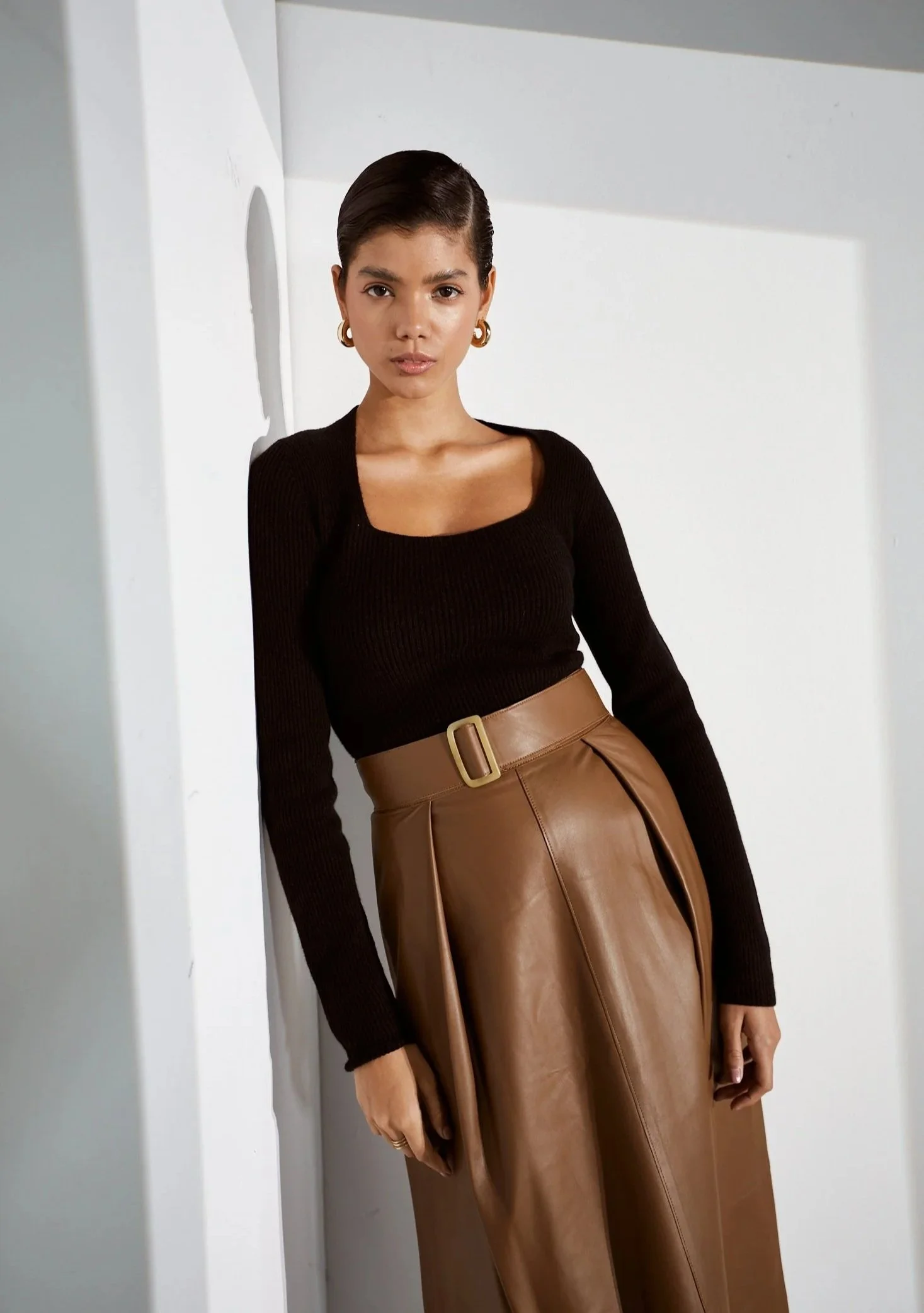 A woman with short dark hair wearing a black long-sleeve top and a brown leather high-waisted skirt with a belt, standing against a white wall, looking at the camera.