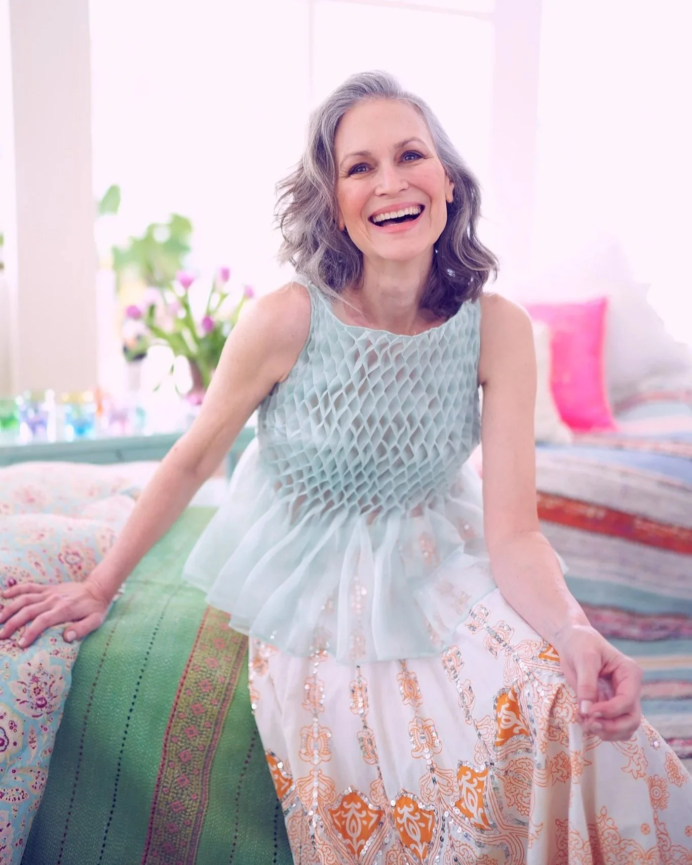An elderly woman with gray hair smiling in a brightly lit room with colorful pillows and flowers in the background.