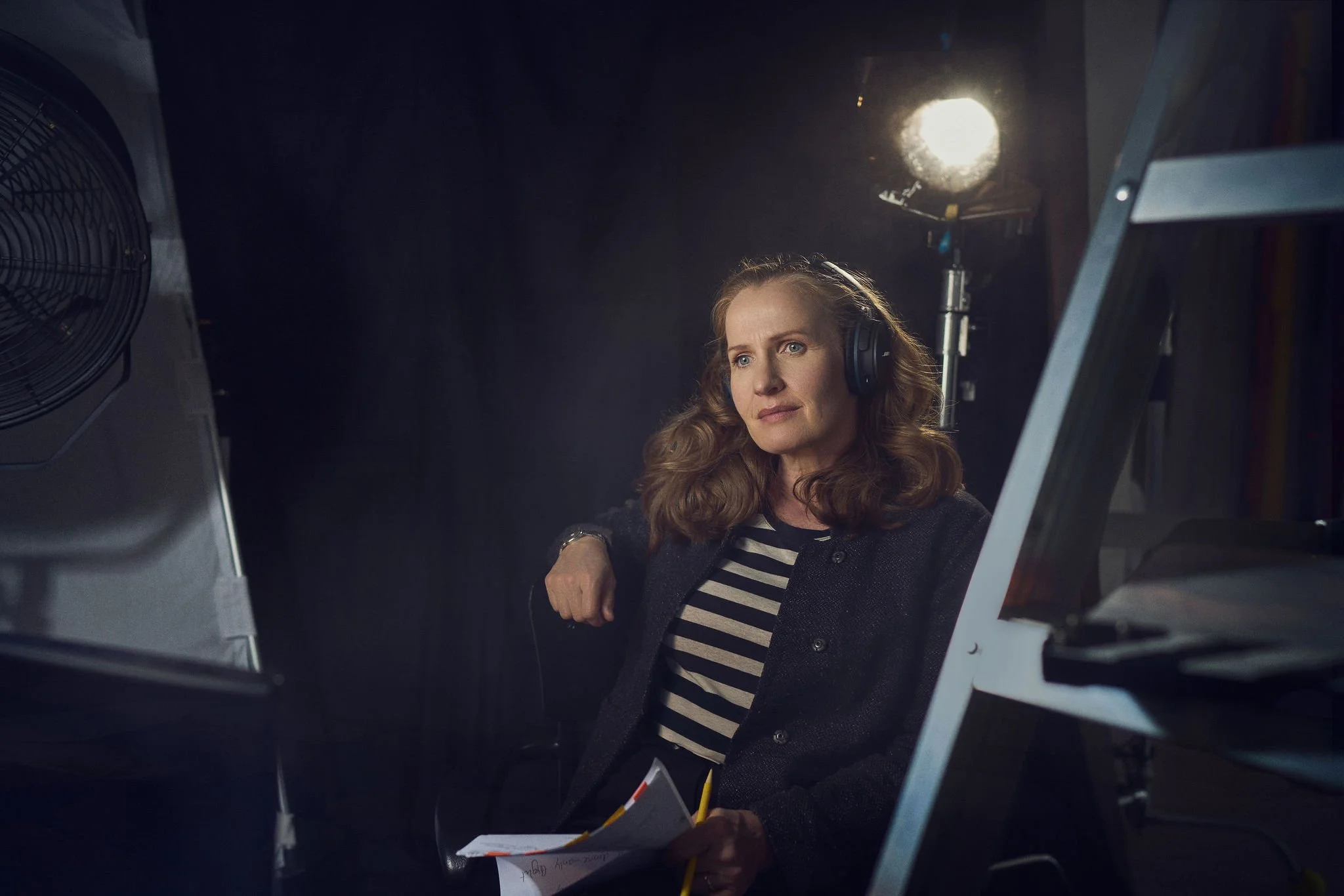 A woman with wavy red hair, wearing headphones, a striped shirt, and a dark jacket, sitting in a studio with lighting equipment and holding papers, appears to be recording or participating in a voice or video project.