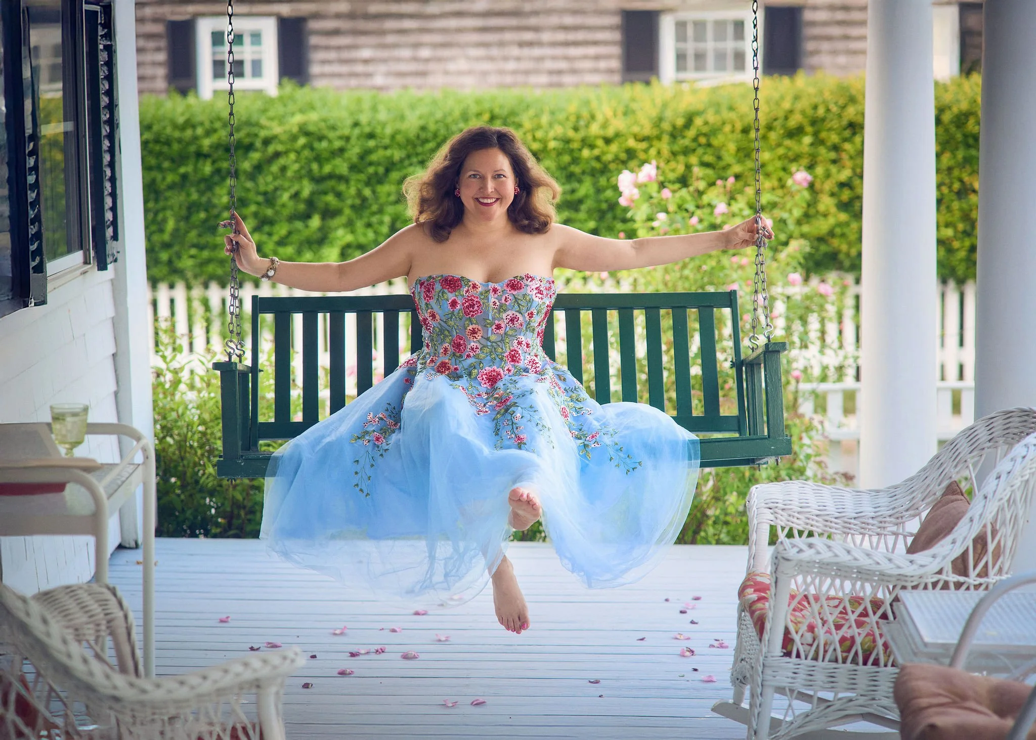 A woman in a floral strapless dress sitting on a porch swing, smiling, with her feet off the ground and surrounded by pink flower petals scattered on the porch.