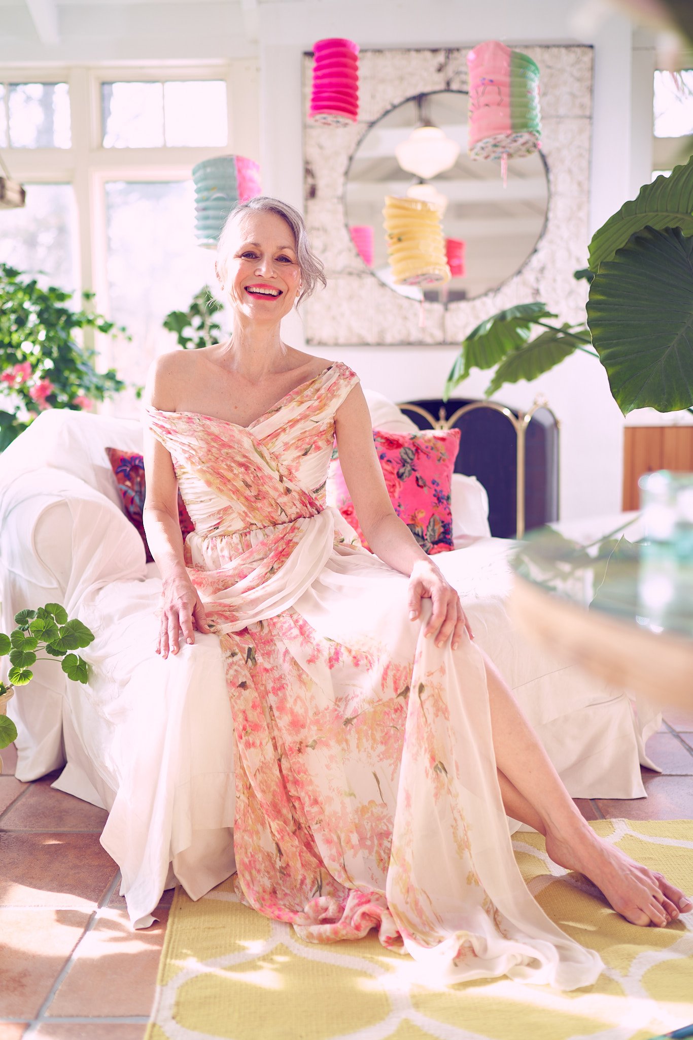 An elderly woman with gray hair smiling while sitting on a white sofa in a well-lit room decorated with colorful paper lanterns and plants.