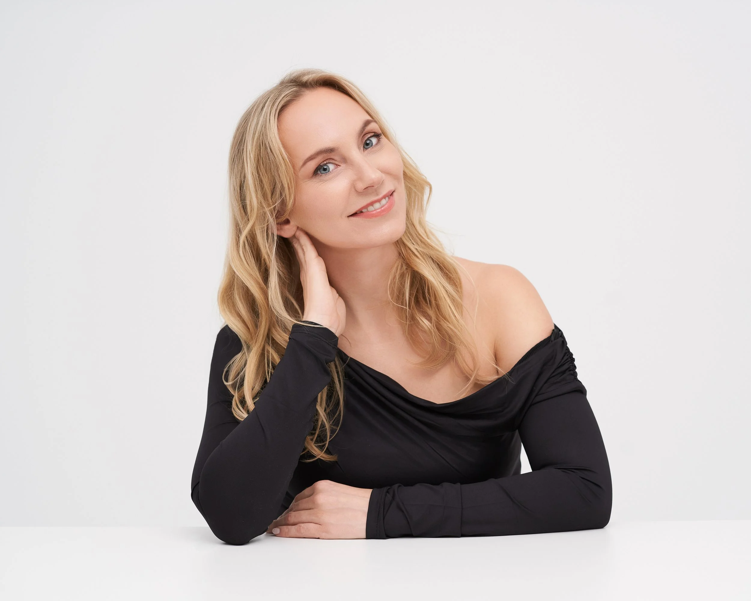 Portrait of a young woman with blonde hair, smiling gently, wearing a black off-shoulder top, with her left hand resting on her cheek and her right arm on a white surface against a plain white background.