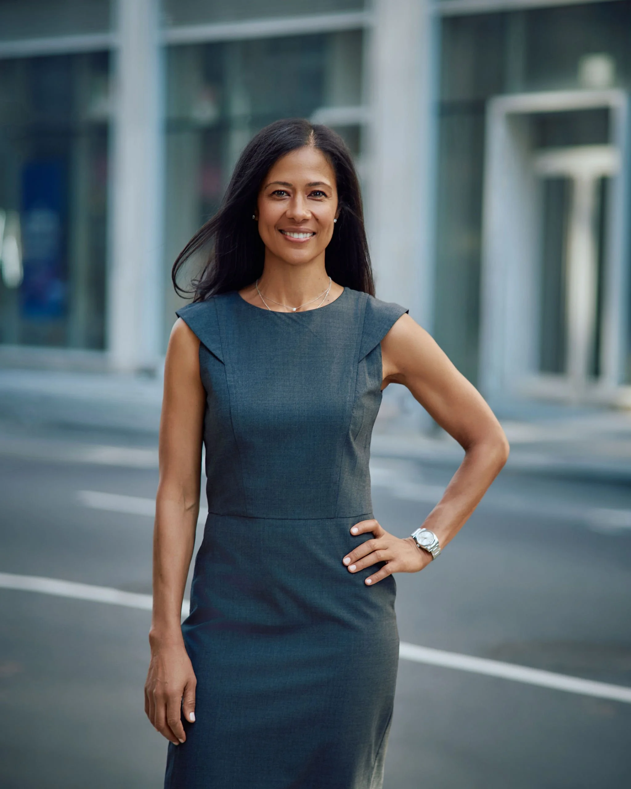A woman in a sleeveless dark gray dress standing outdoors in an urban area, smiling at the camera with hand on her hip.
