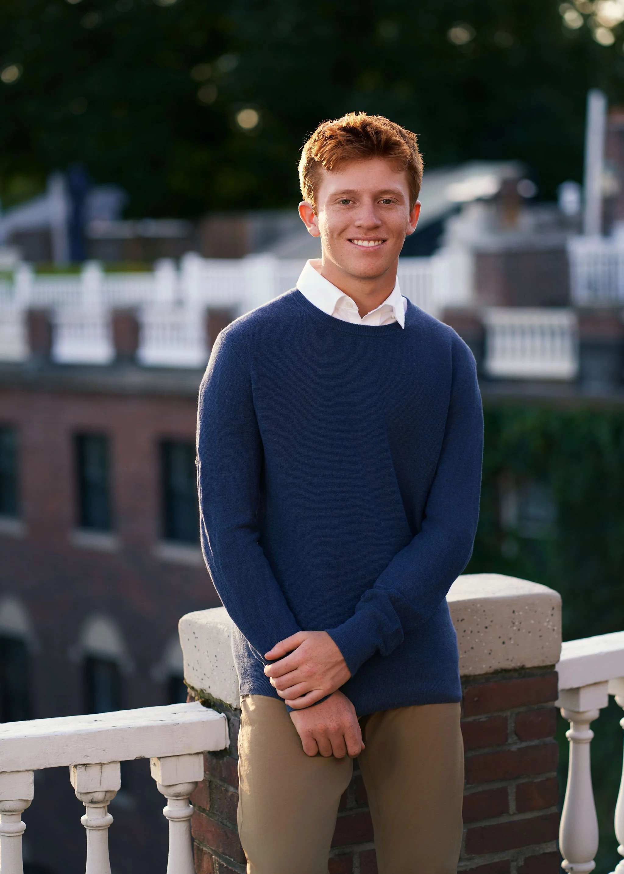 A young man with red hair wearing a white shirt, navy sweater, and khaki pants, smiling and standing outdoors by a white railing with a blurred background of buildings and trees.