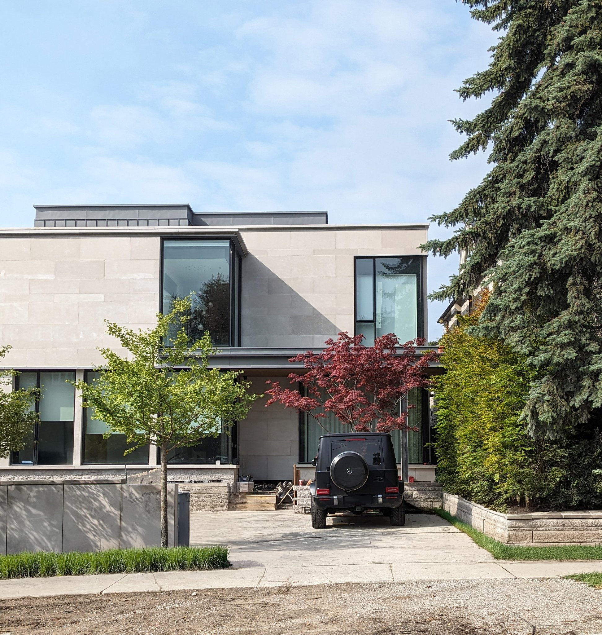 Modern multi-story house with large glass windows, surrounded by trees, with a dark vehicle parked in front, and a partly cloudy sky overhead.