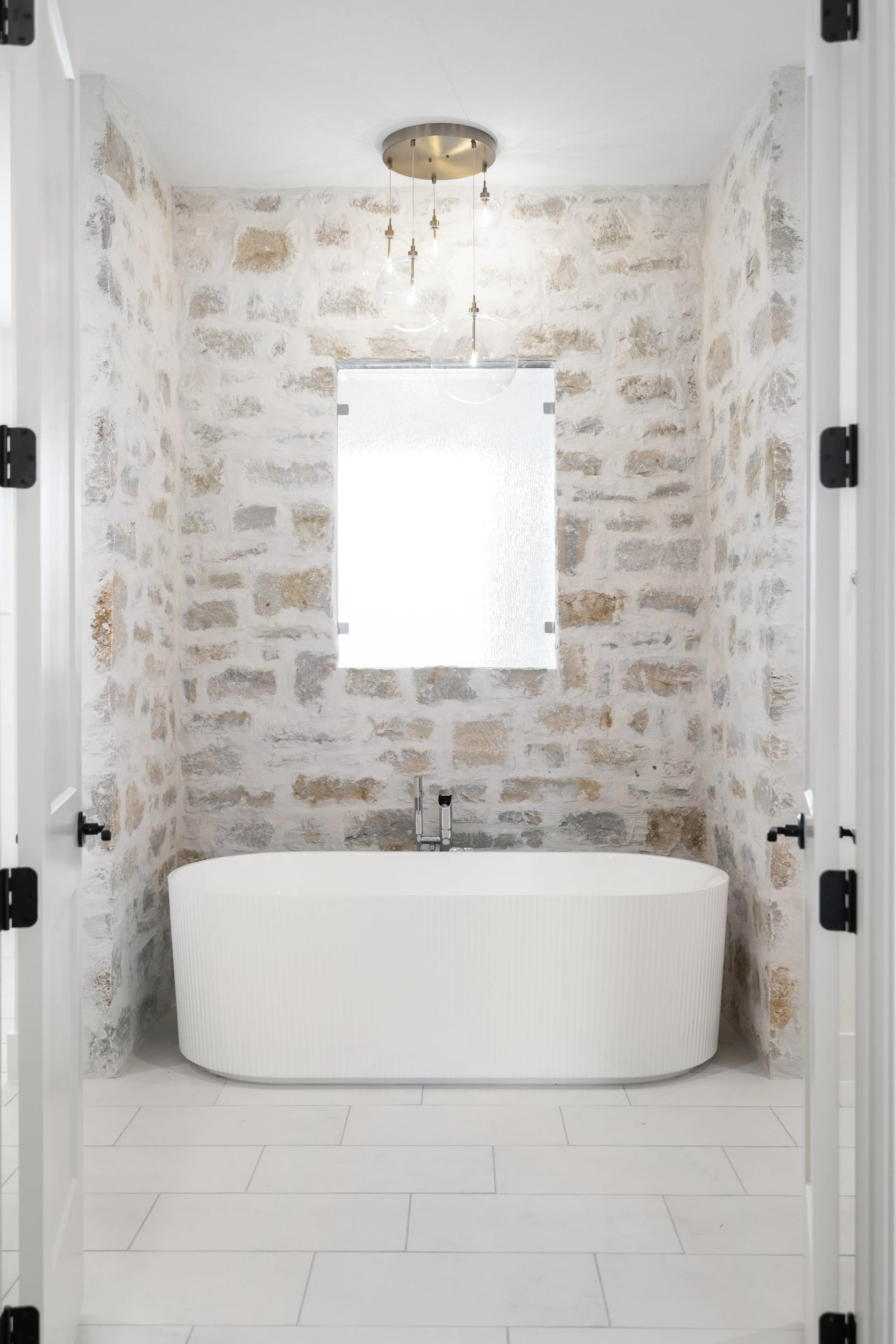 Modern bathroom with a freestanding bathtub, exposed brick walls, a window, and a unique chandelier lighting fixture.