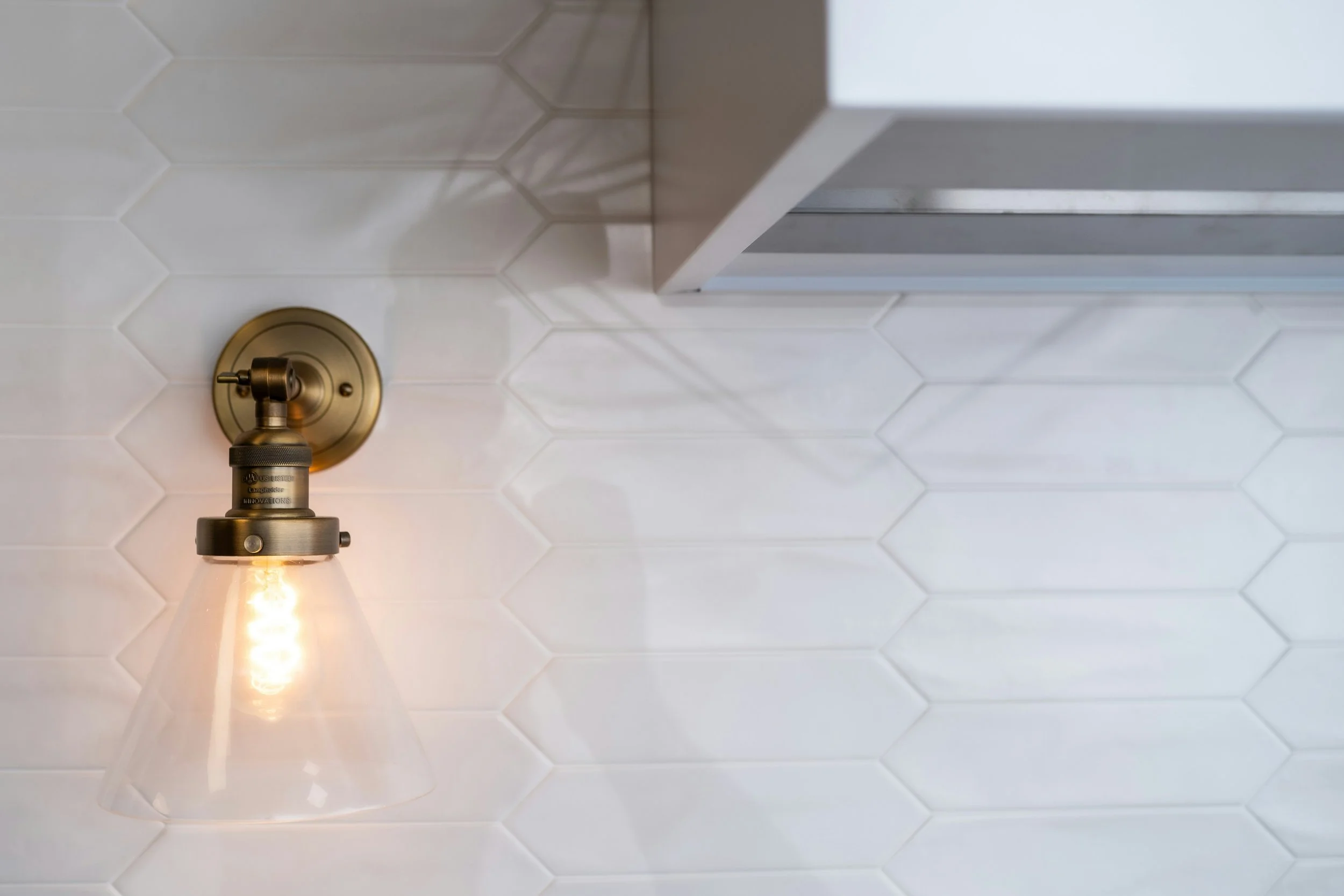 Close-up view of a wall-mounted industrial-style light fixture with a clear glass shade and visible filament bulb, set against a white subway tile wall.