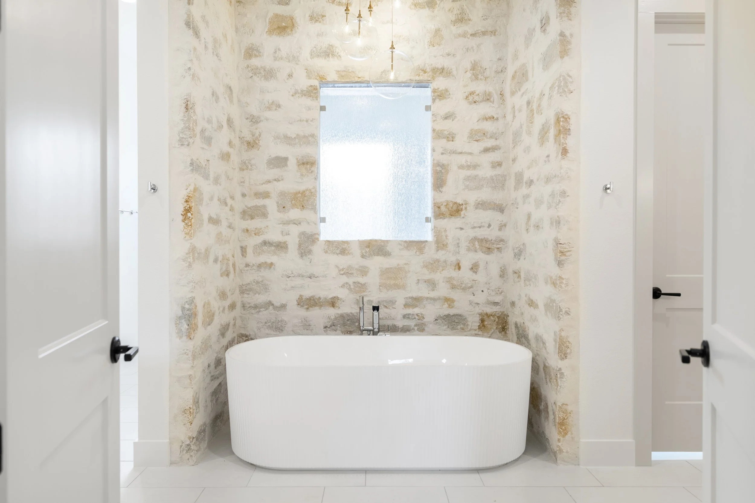 Modern bathroom with a freestanding bathtub against a textured brick wall, a small frosted window above, and white doors on either side.