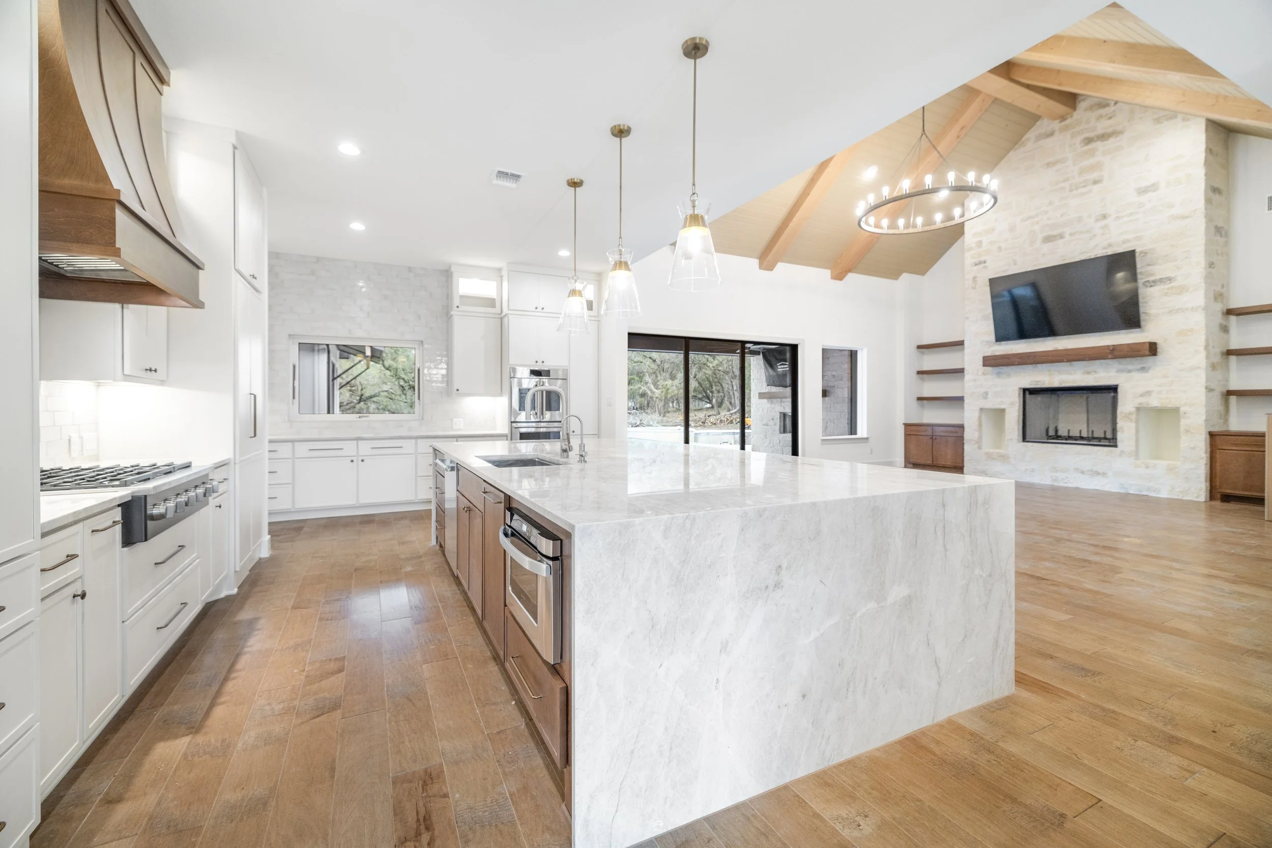 Modern open-concept kitchen and living room with wooden floors, white cabinets, marble island, stone fireplace, large sliding glass doors, and vaulted ceiling with wood beams.