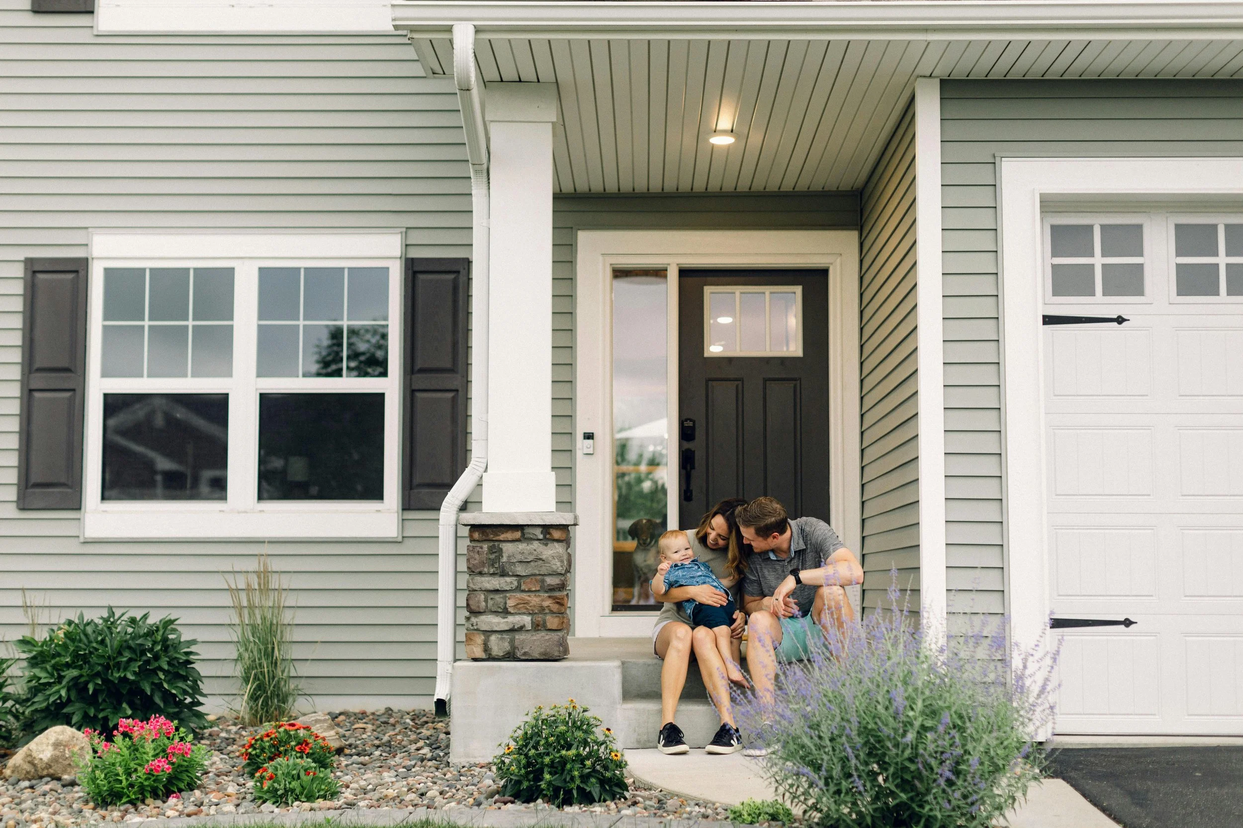 A family of three sitting on the front steps of a house, with a woman, a man, and a young child, smiling and enjoying time together outside their home.